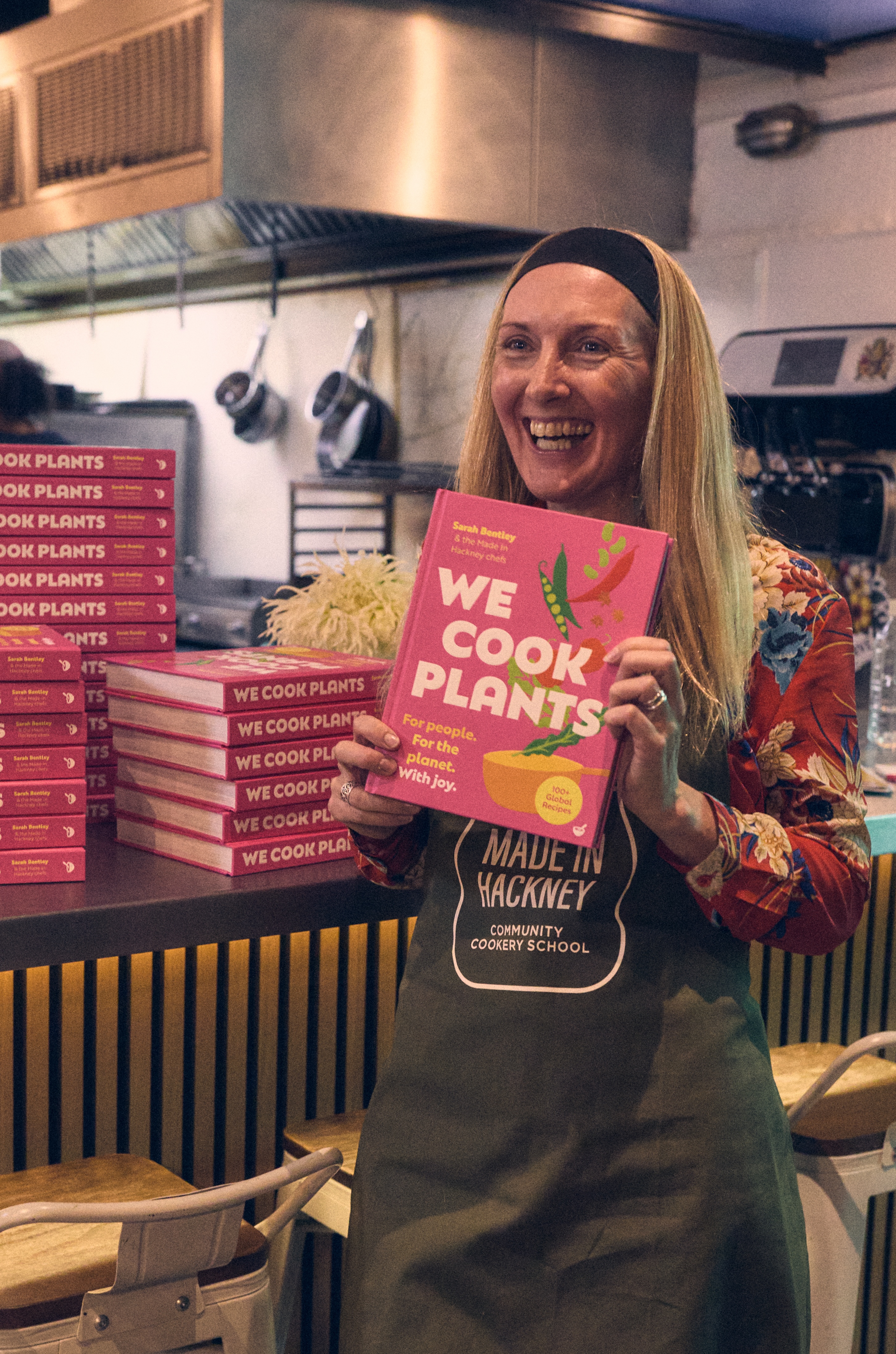 Blonde woman in apron holds "We Cook Plants" cookbook in commercial kitchen with stack of identical books on counter behind her.