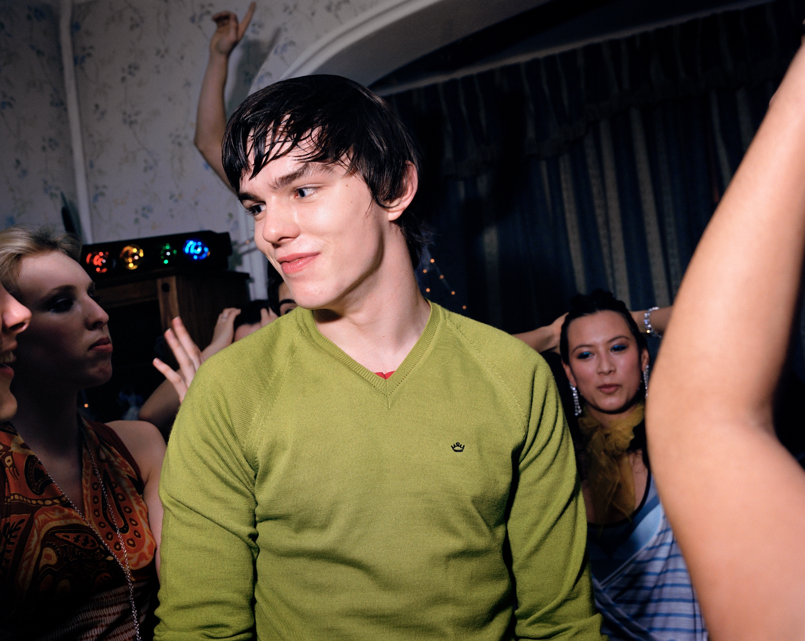 A young man with short, dark hair wearing a green v-neck jumper, surrounded by other people in a dimly lit room.