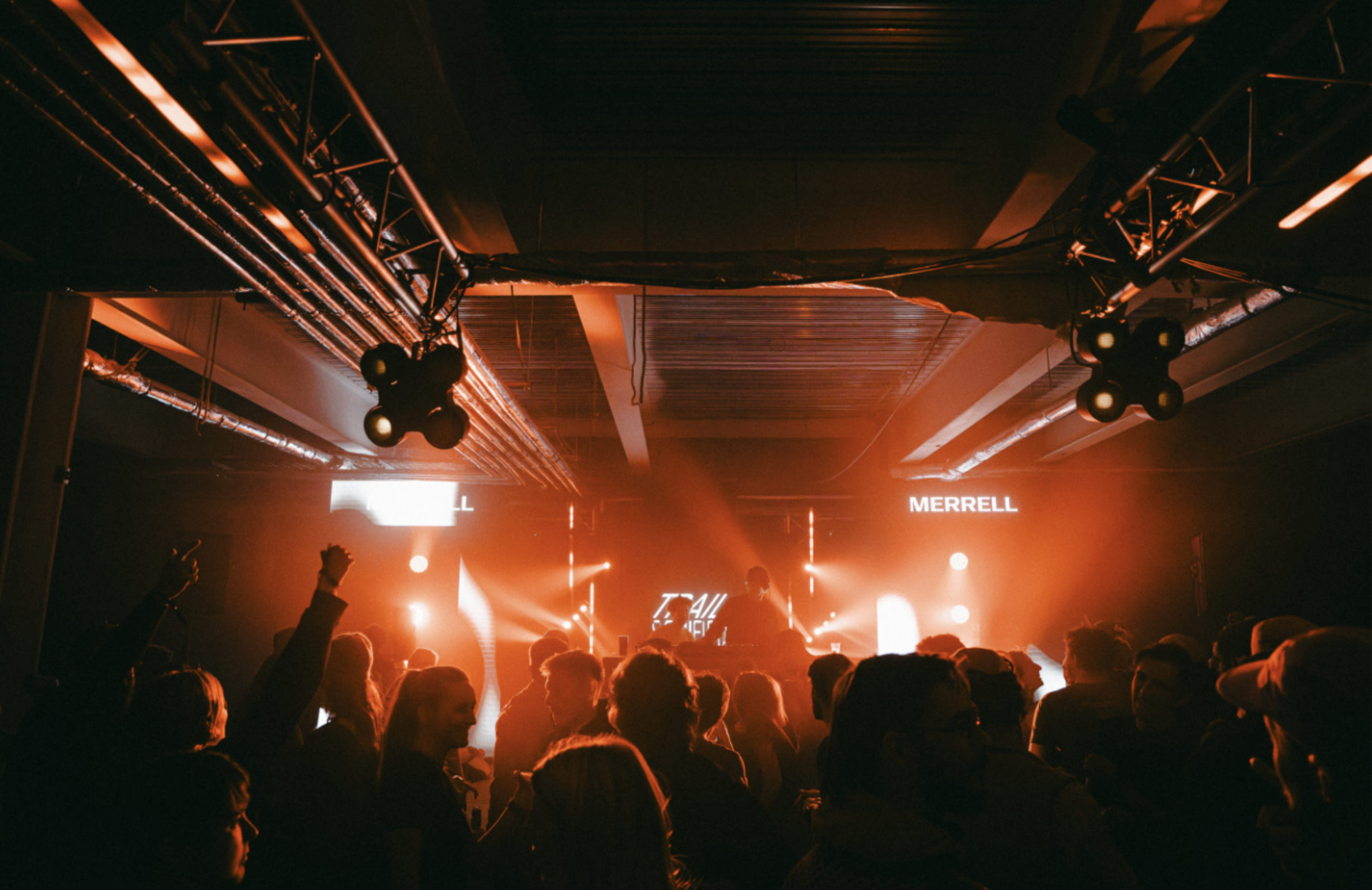 Concert venue with crowd silhouettes, orange stage lighting, exposed ceiling beams, and "MERRELL" sign visible on back wall.
