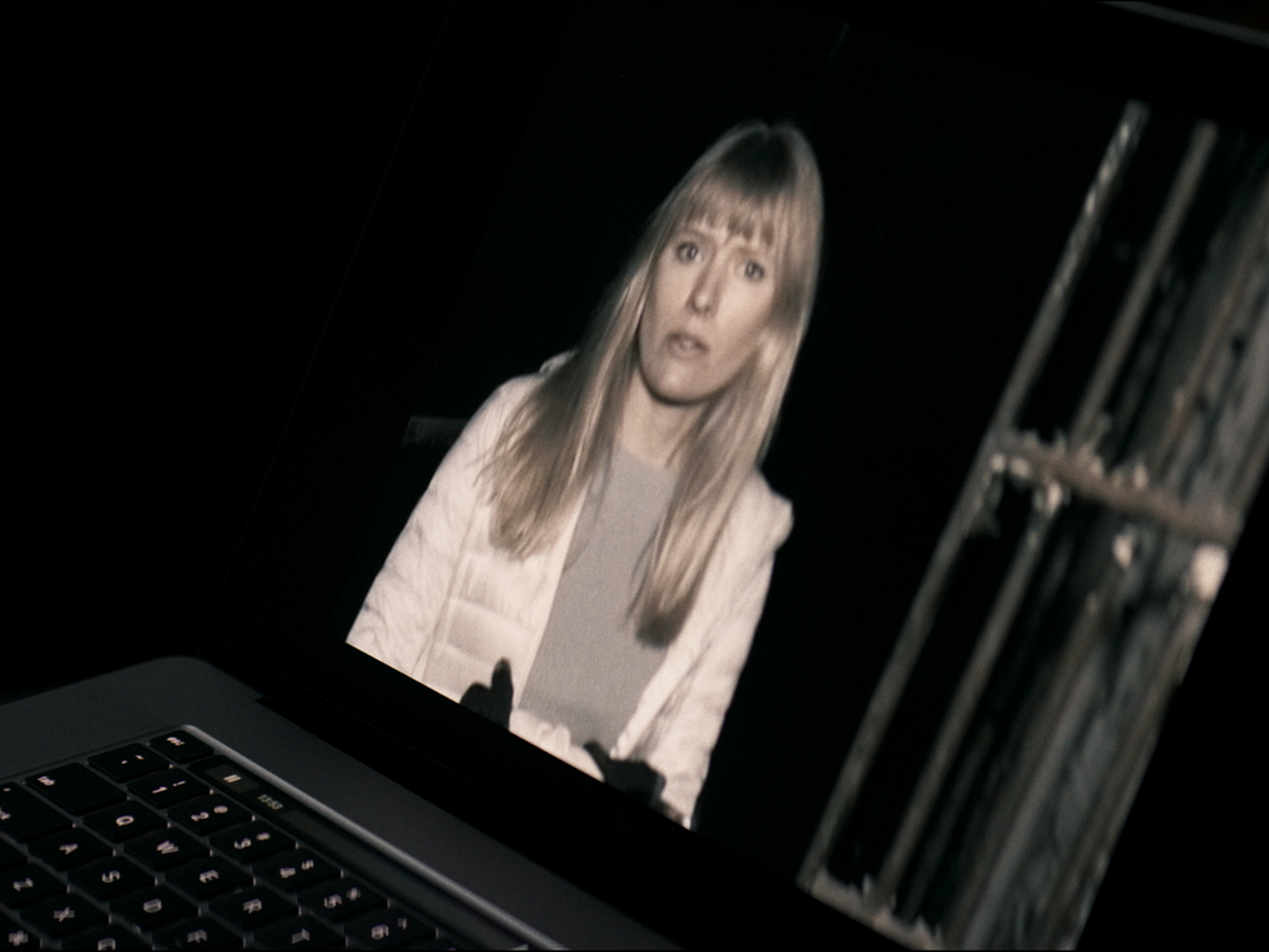 Blonde woman with fringe displayed on laptop screen, second laptop visible to the right, dark background.
