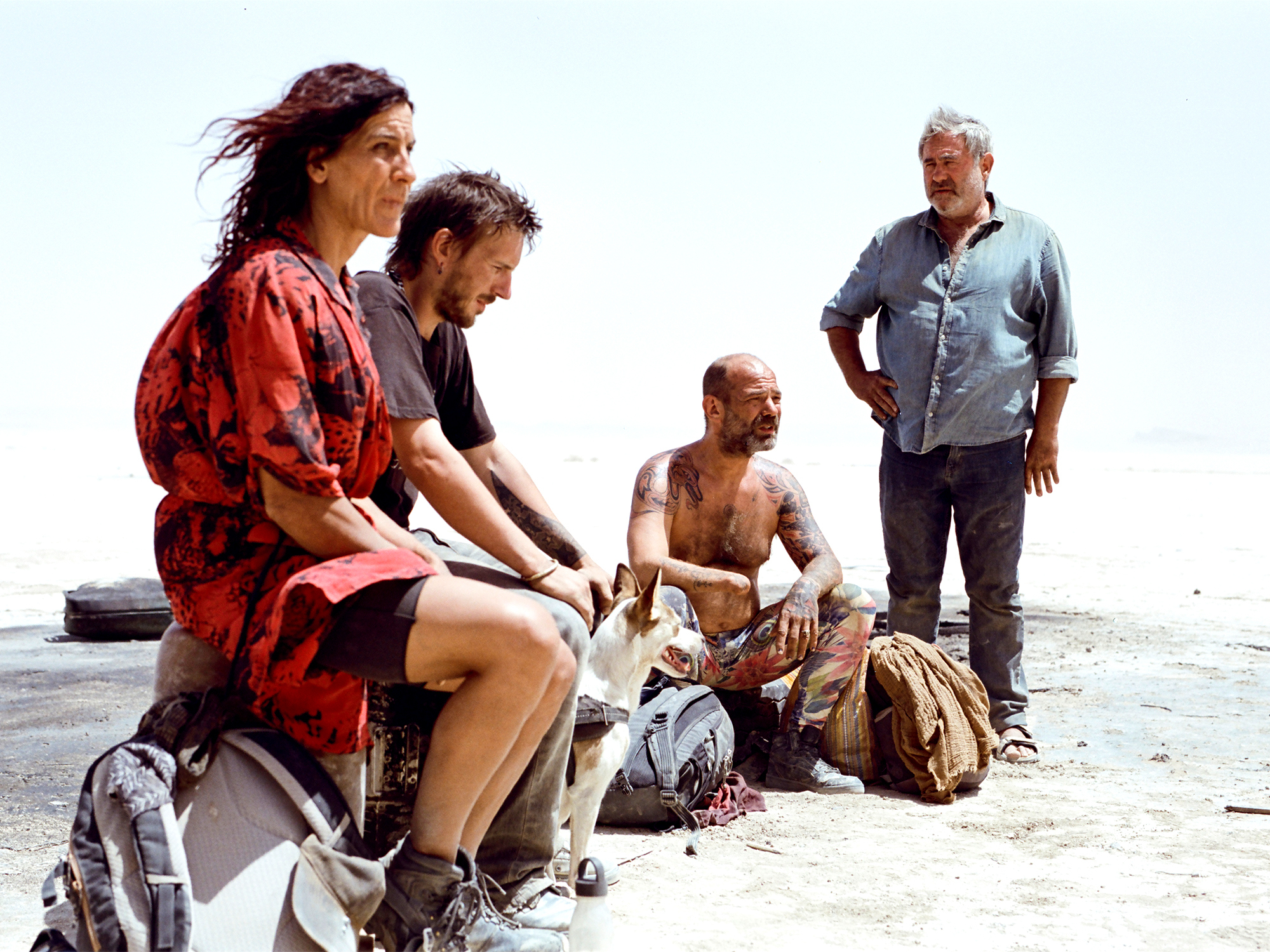 A group of four adults, three men and one woman, sitting on a beach. The woman wears a bright red patterned dress, while the men wear more casual clothing. The scene appears to capture a casual, relaxed moment.