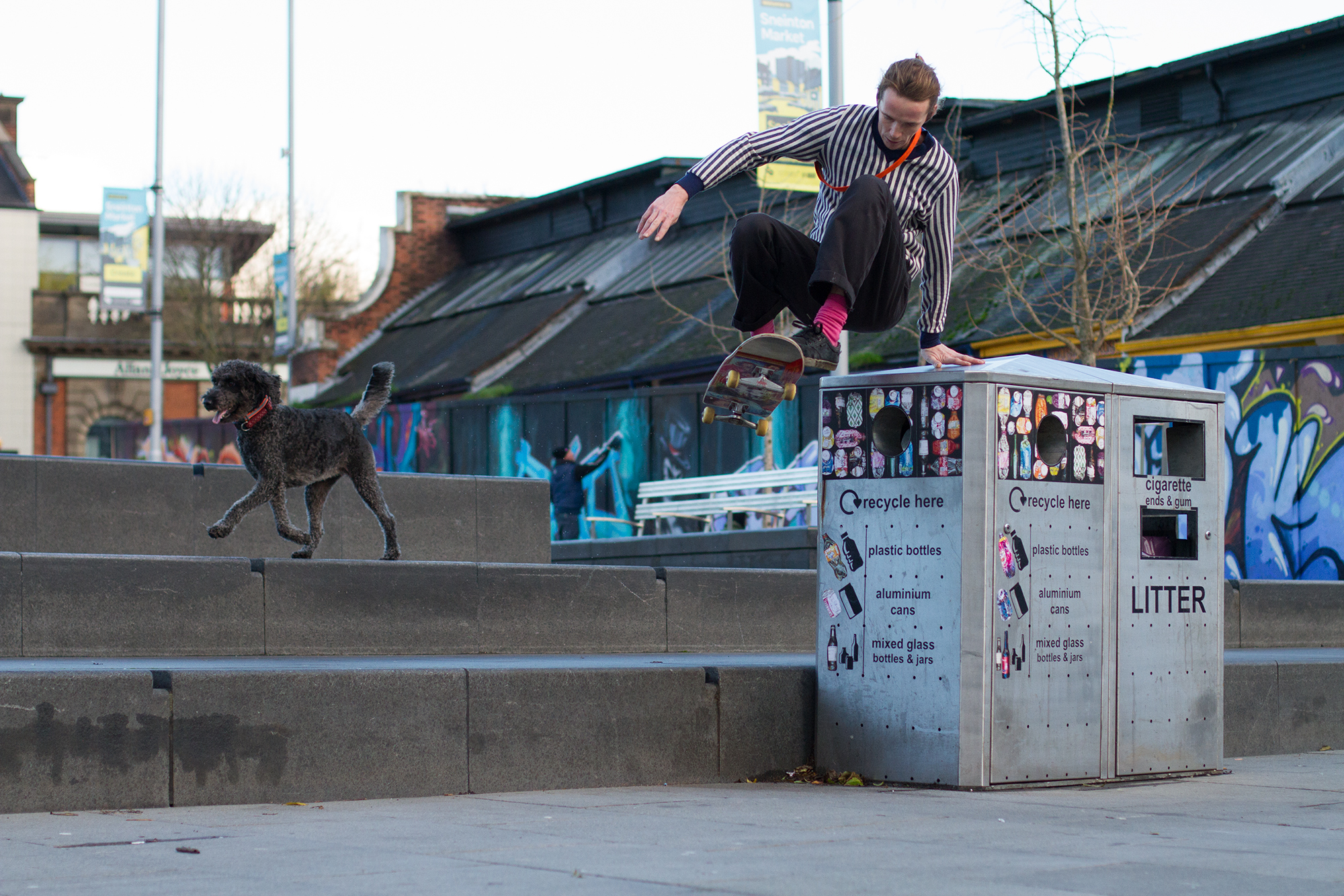 Man in striped shirt crouching on concrete ledge near black dog and recycling bins, with graffitied wall behind.