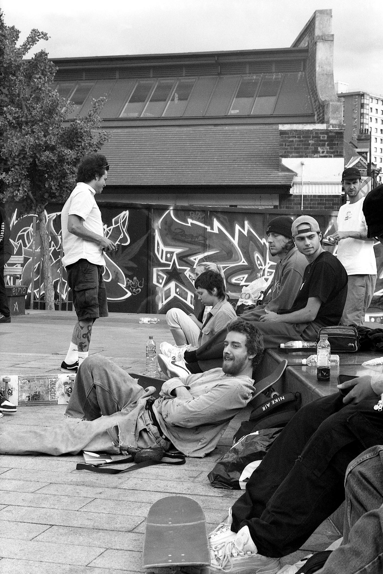 Group of young people gathered in urban plaza, some seated on ground, one standing, with graffitied wall and buildings behind.