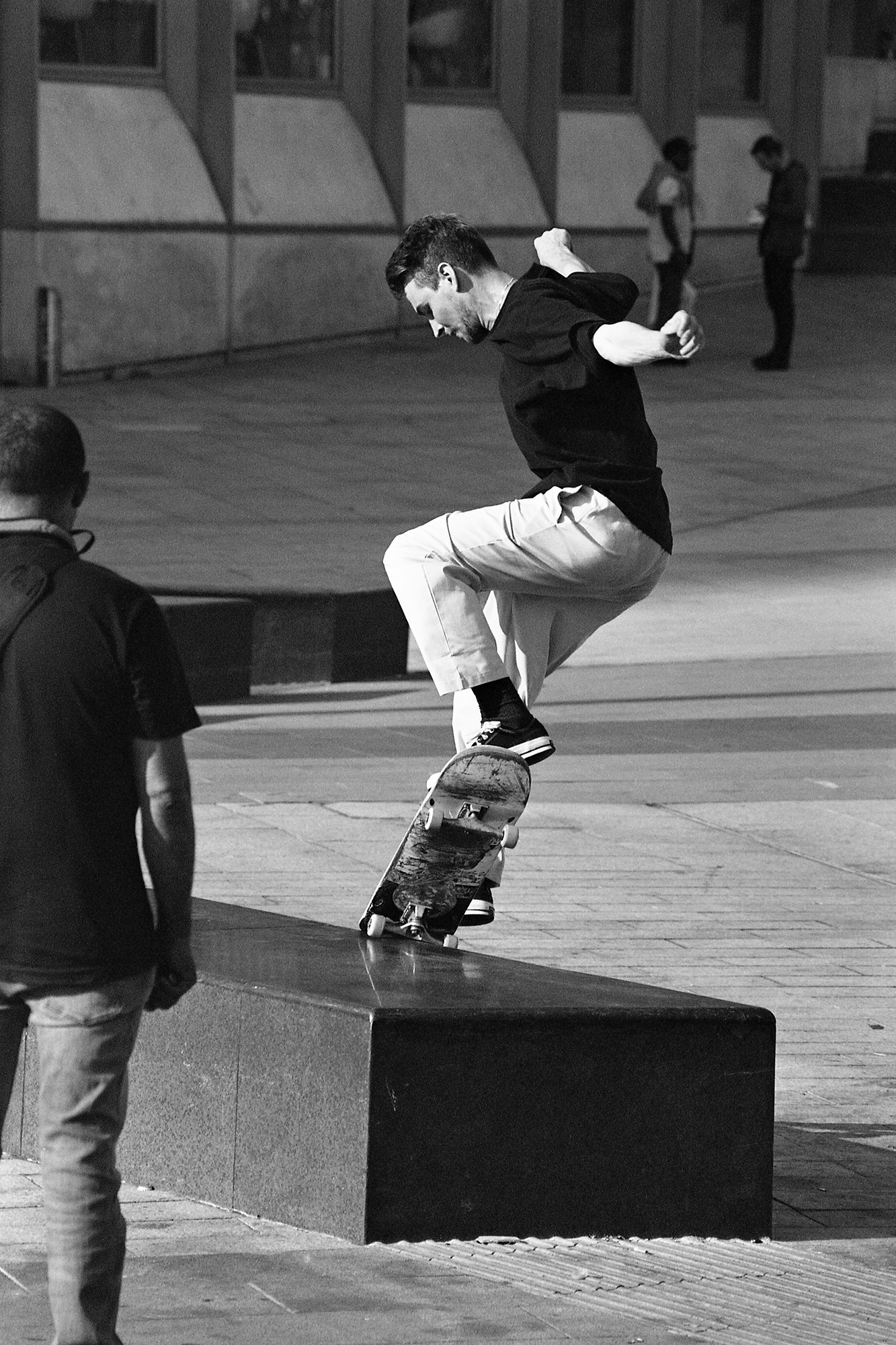 Skateboarder performing trick on concrete ledge in urban plaza, black and white image with building in background.