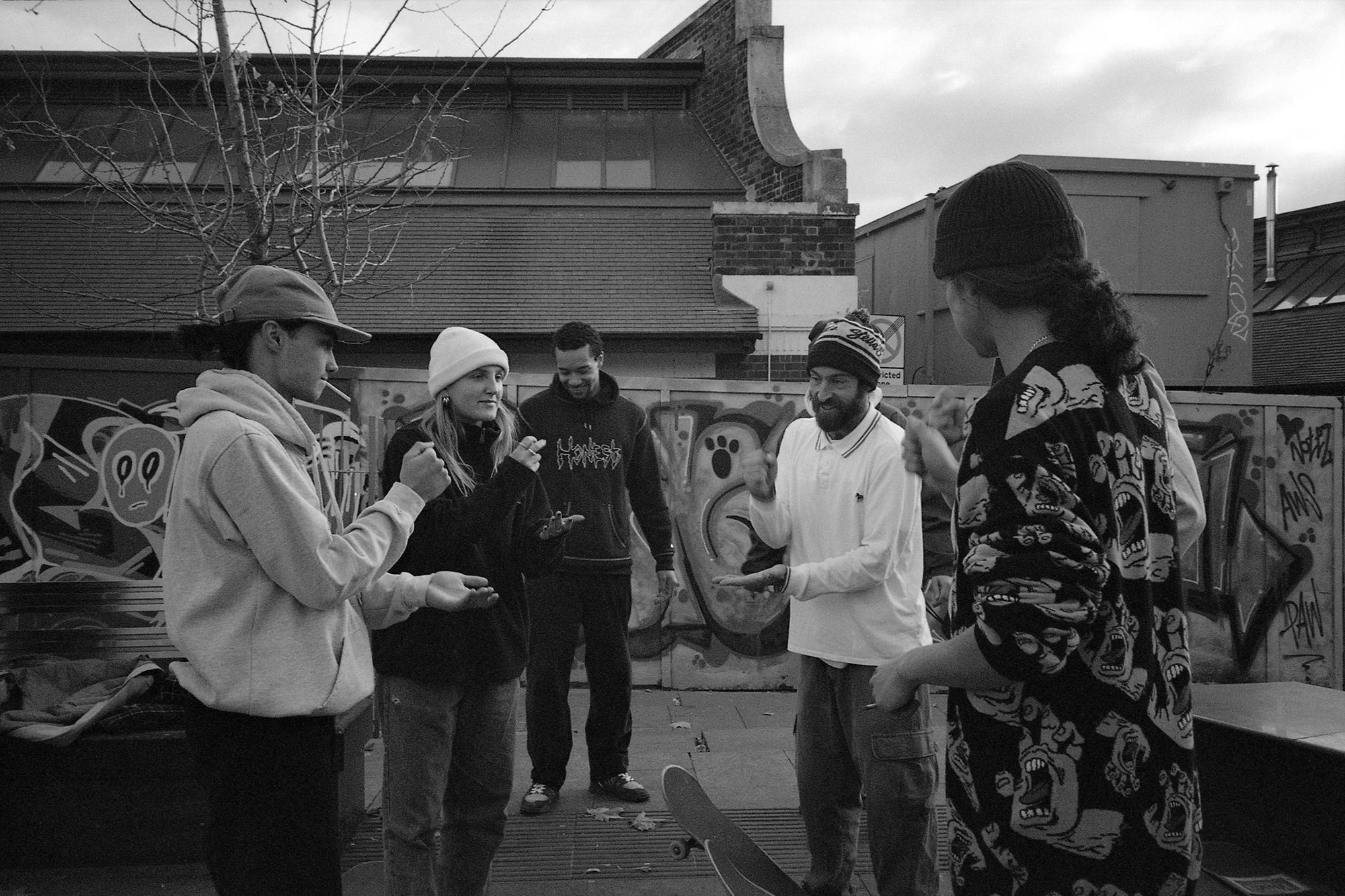 Black and white image of five people standing in urban setting with graffitied wall, residential buildings and bare tree in background.