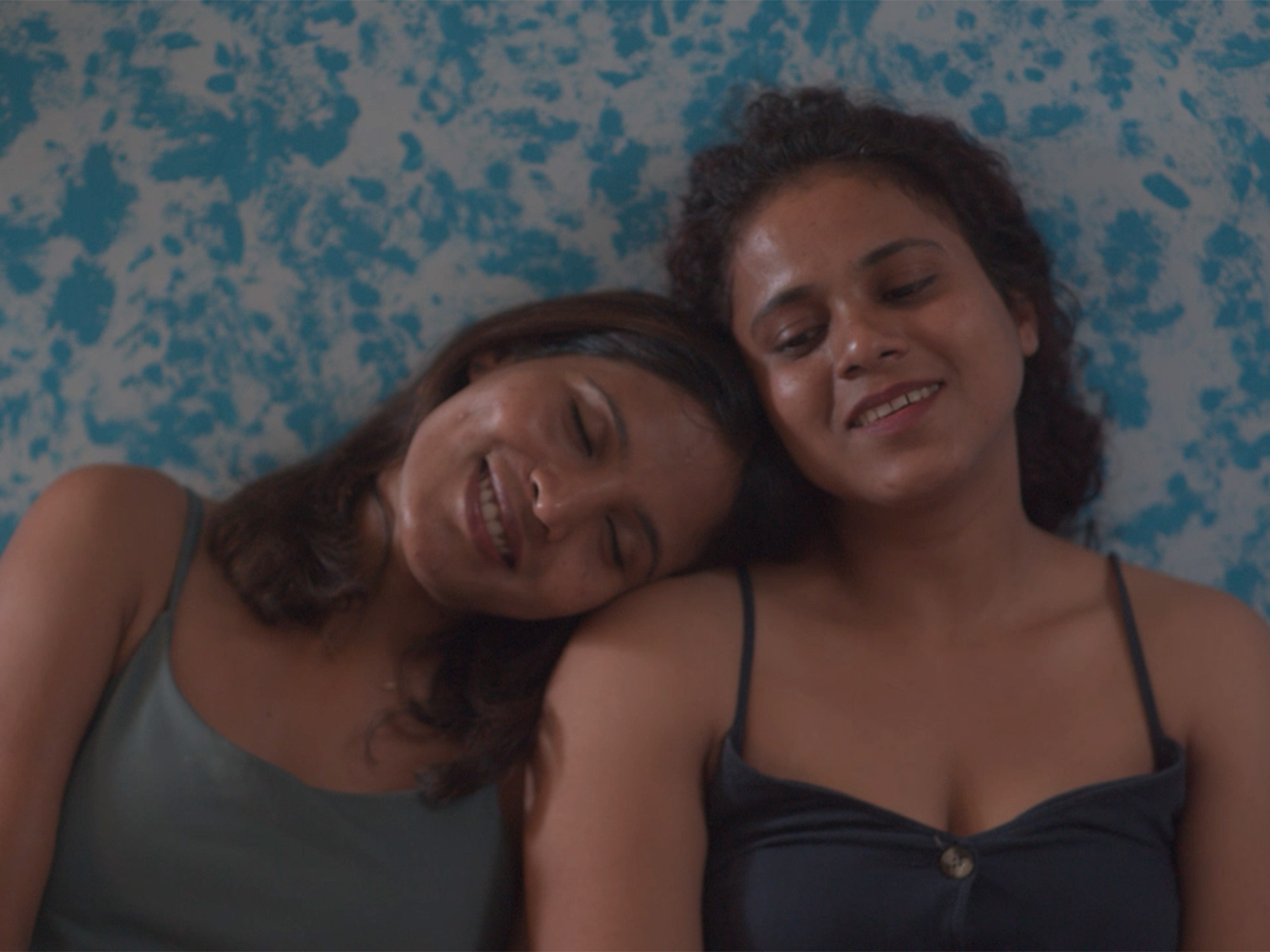Two women lying down, one resting head on the other's shoulder. Blue mosaic tile background. Both wearing swimwear or sleeveless tops.