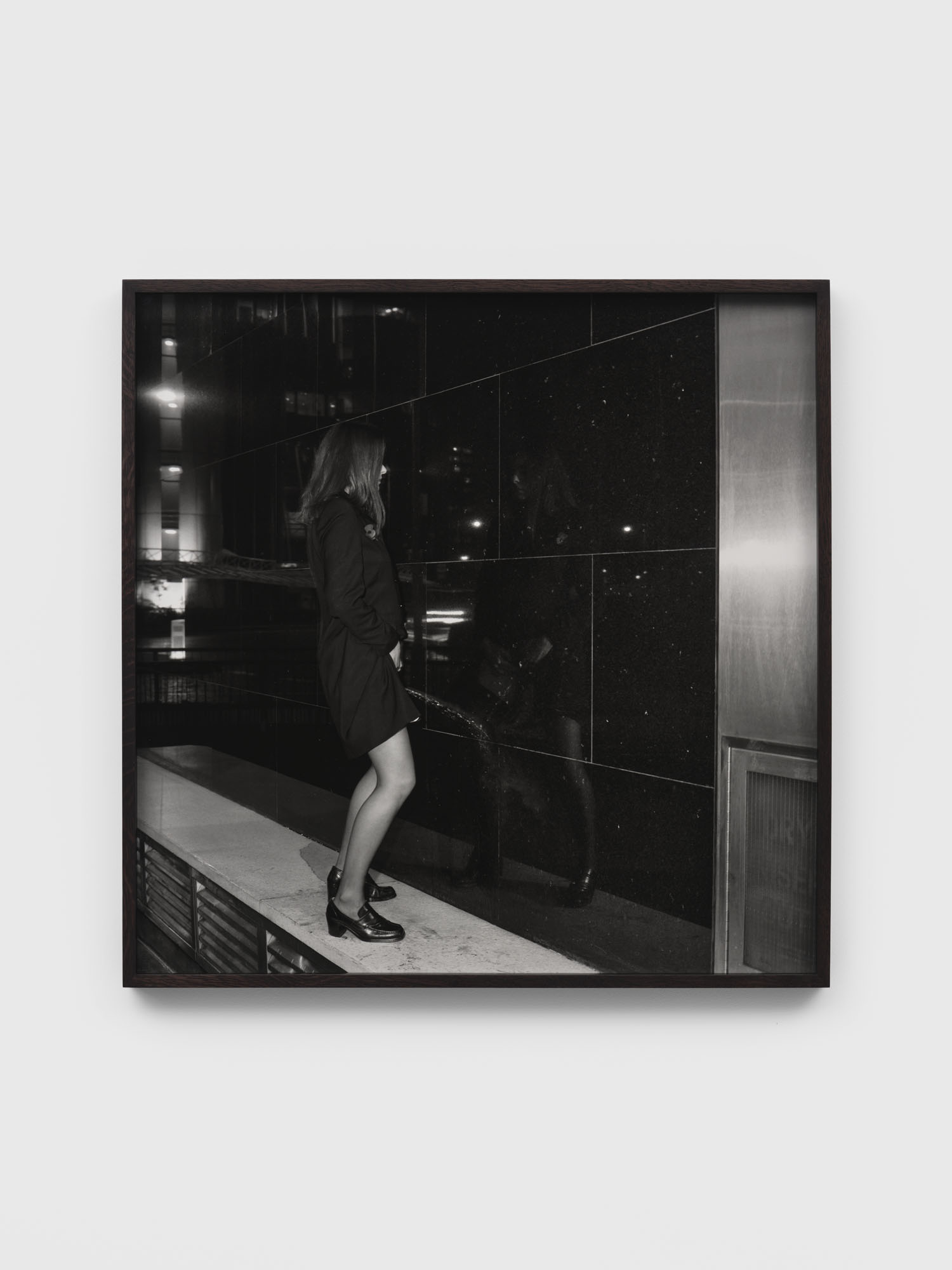 Black and white photograph in dark frame showing person in kitchen with reflective surfaces and dramatic lighting contrasts.
