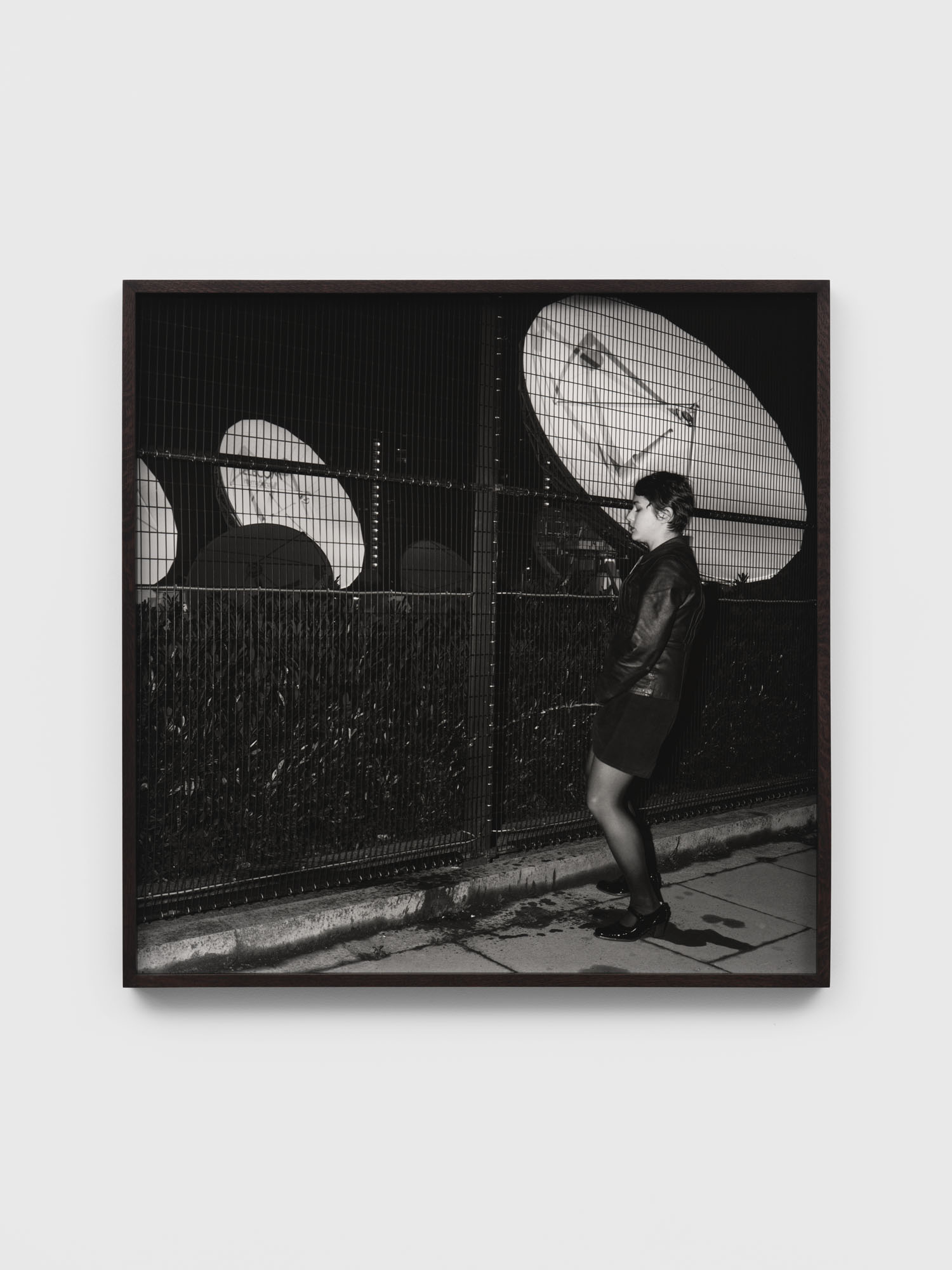 Black and white photograph of person in dark clothing walking past illuminated circular windows at night, mounted in black frame.
