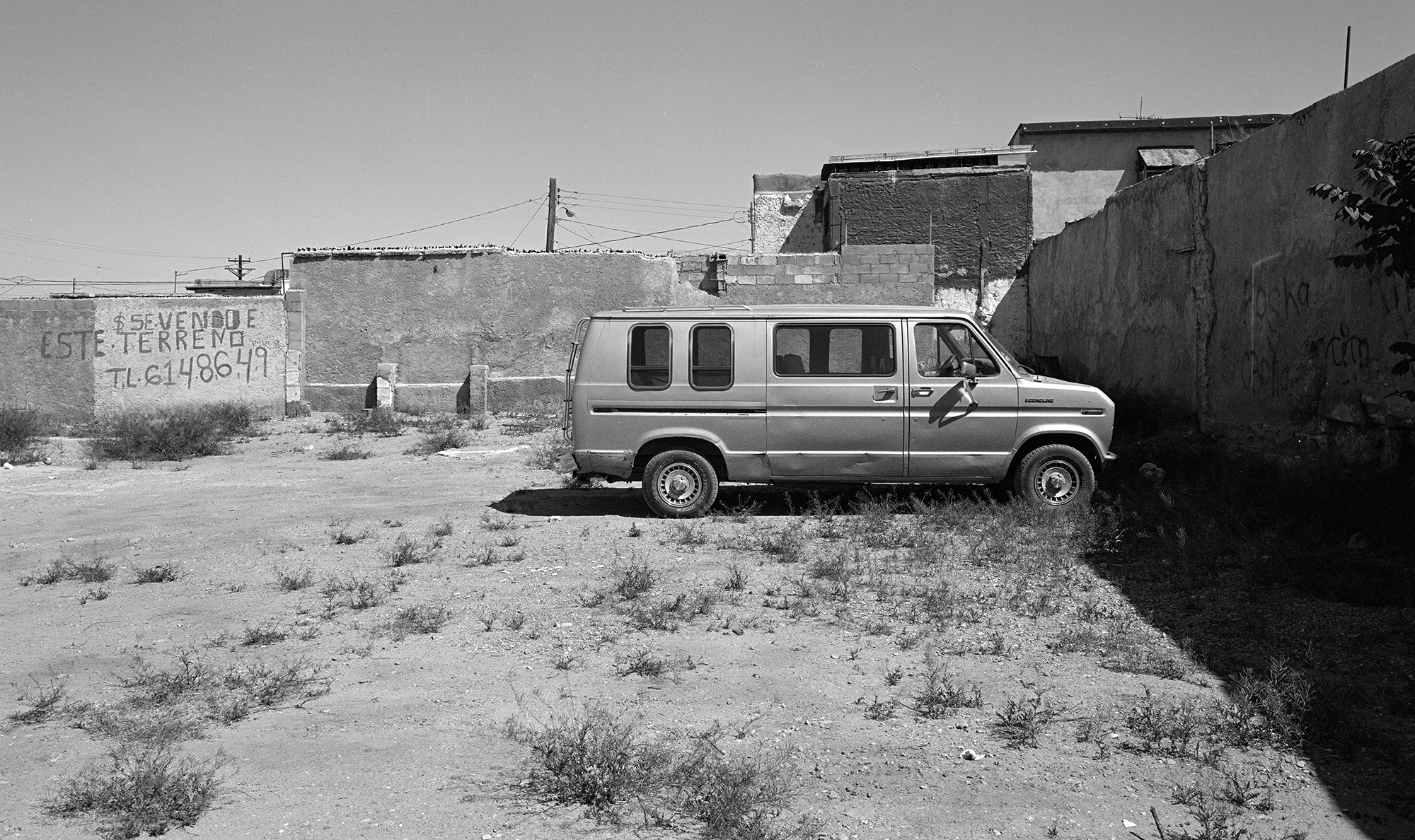 Van parked in dusty courtyard surrounded by concrete block buildings in black and white.