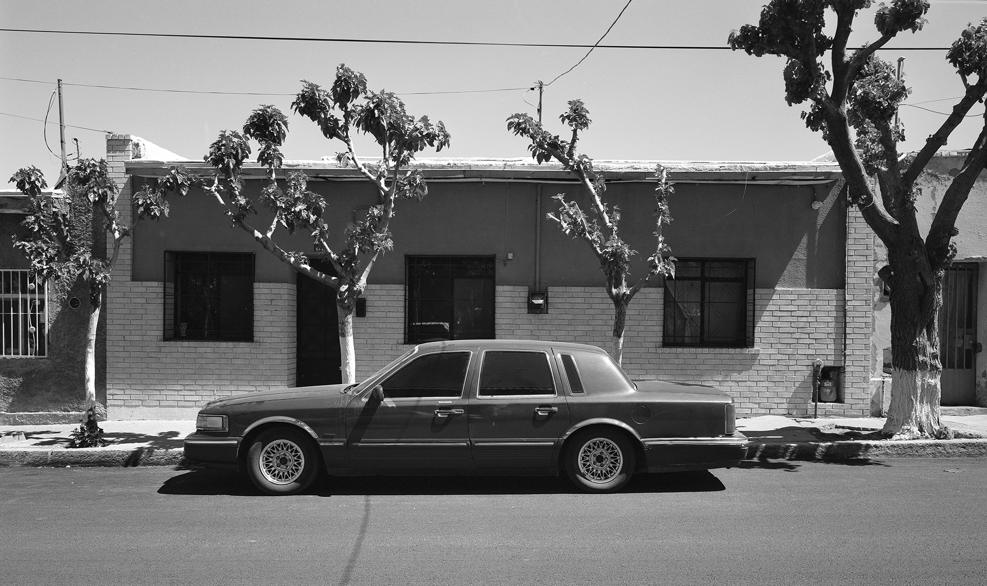 Black and white image of vintage saloon car parked outside single-storey brick building with three windows and bare trees.