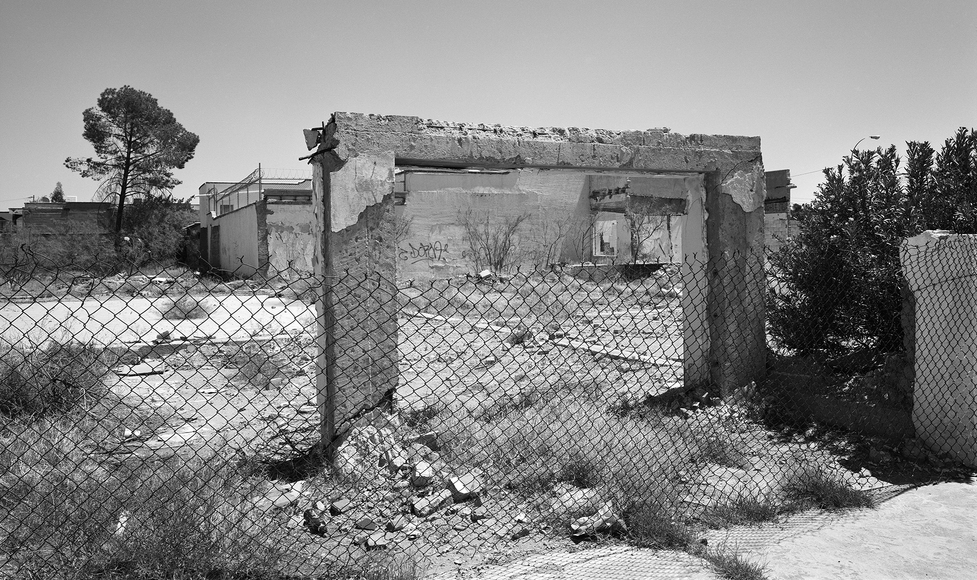 Black and white image of damaged concrete building behind chain-link fence, with trees and cloudy sky in background.