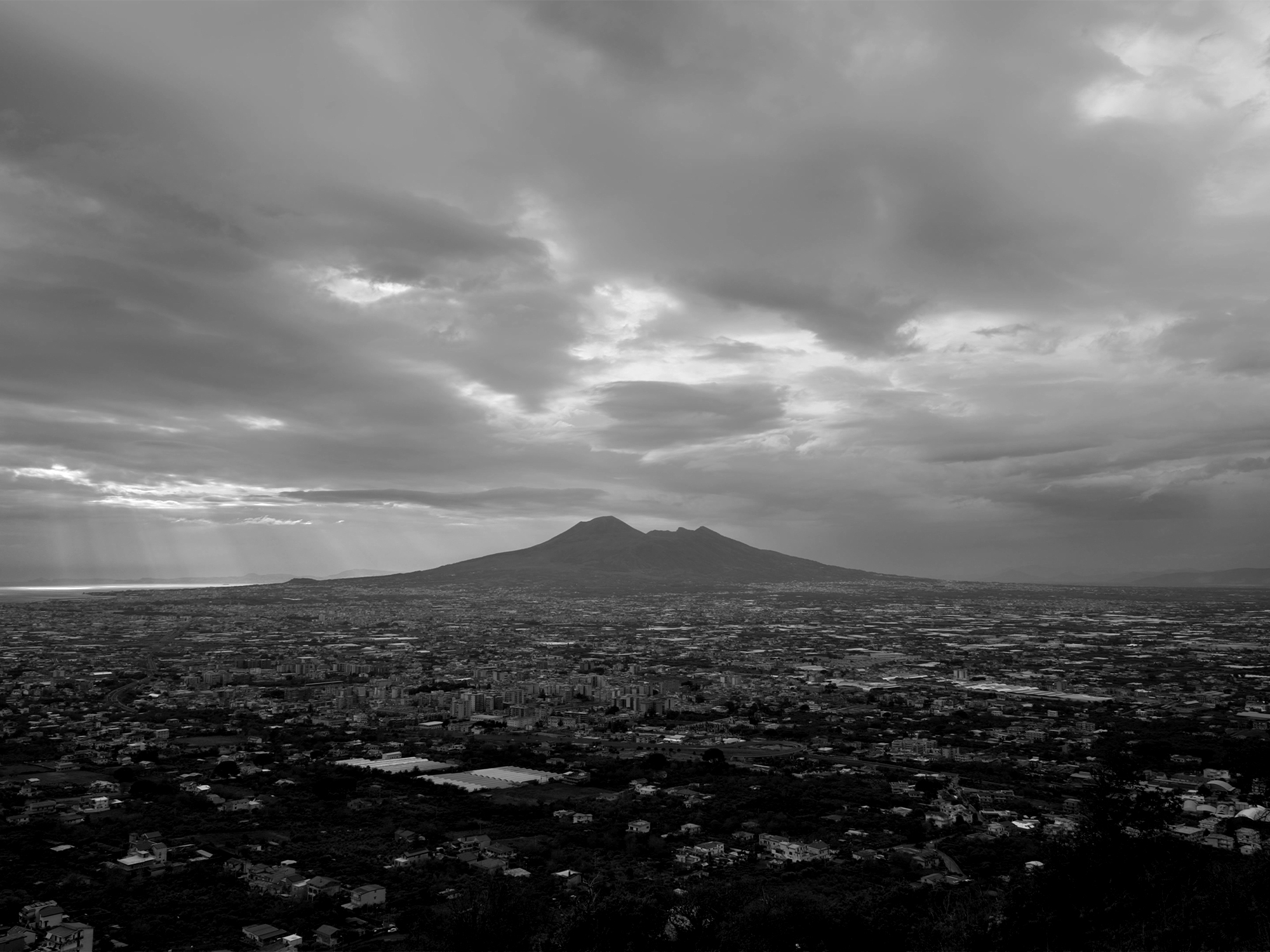 Black and white aerial view of urban sprawl with mountain silhouette in distance under dramatic cloudy sky.