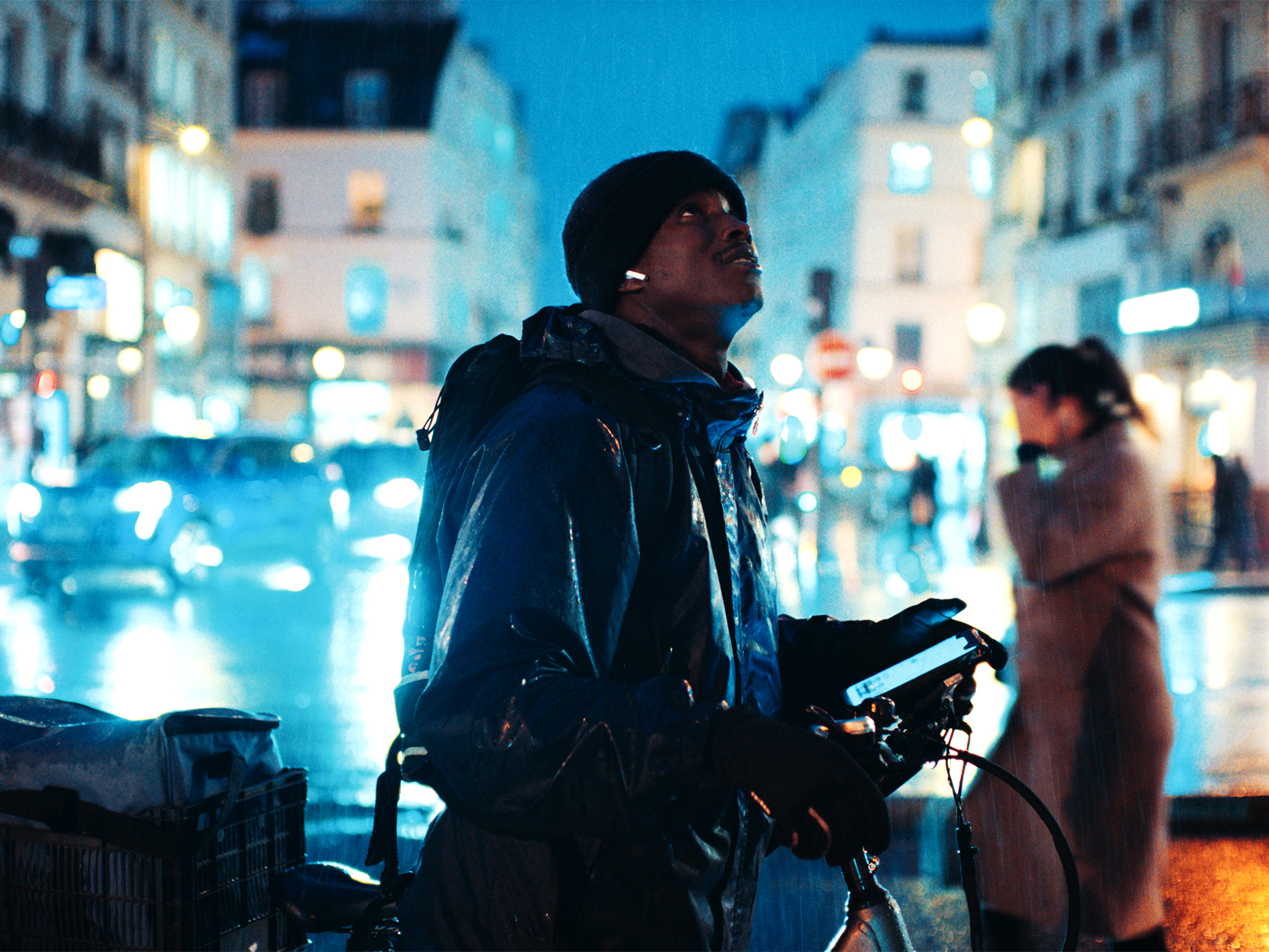 Person in dark jacket and beanie looking upward on city street at night, with blue neon lights and blurred figures in background.