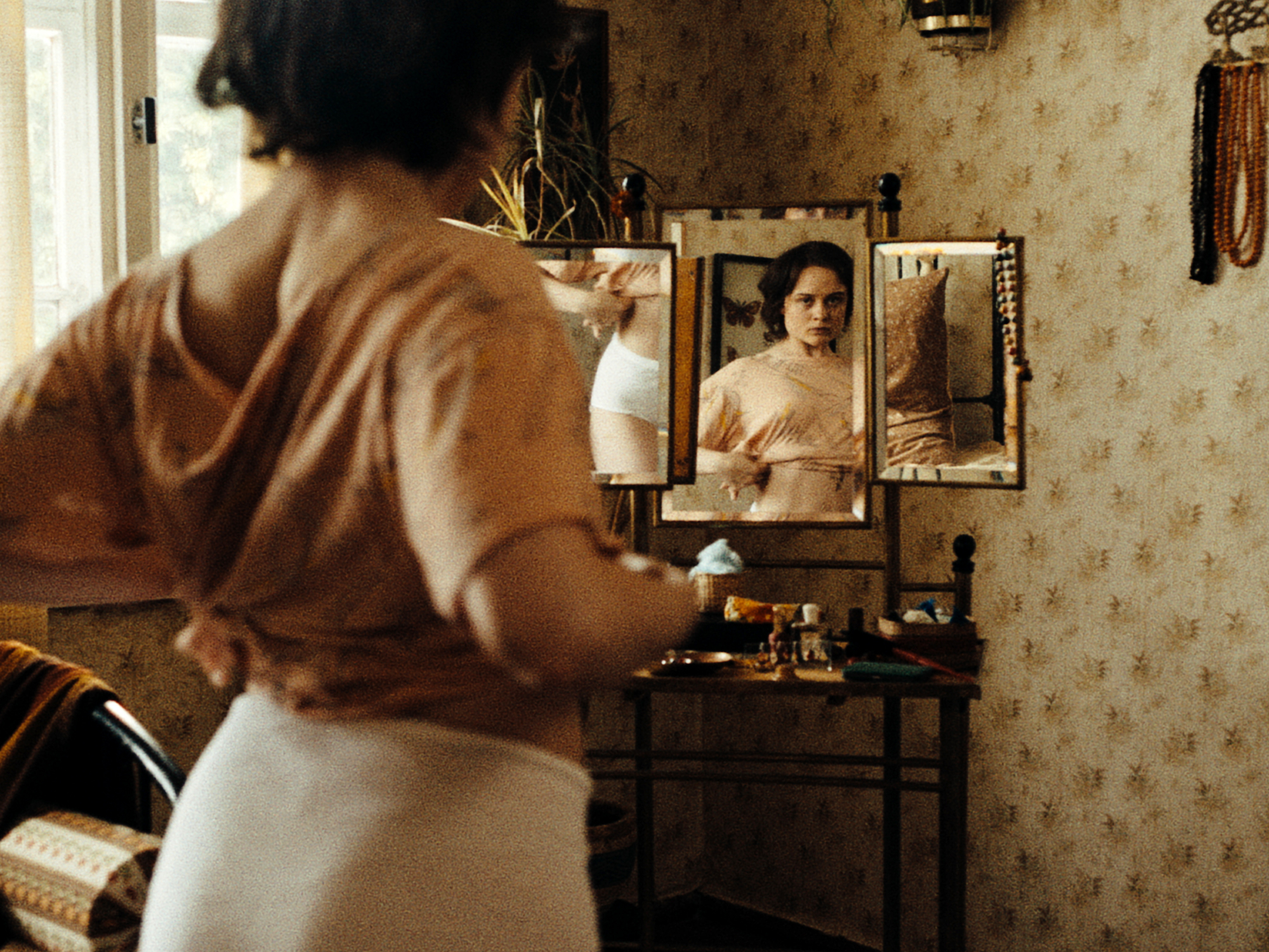 A woman in a brown robe sits at a dresser in a vintage-style room, with framed portraits on the wall behind her.