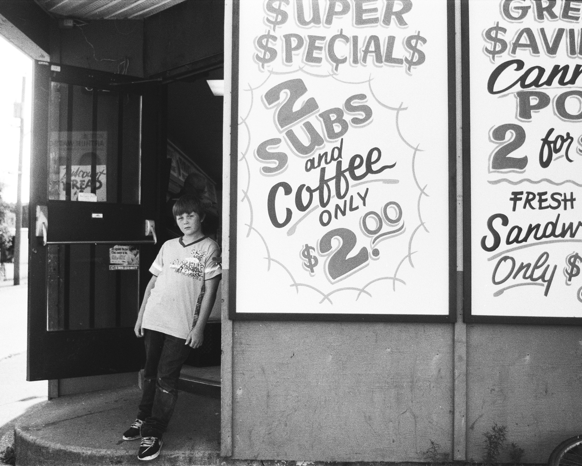 Black and white image of a storefront with signs advertising "Super Specials" and "Great Savings". A person is visible standing in the doorway.