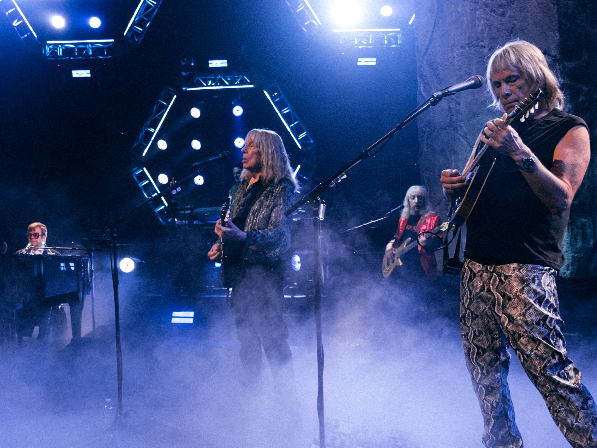 Rock band performing on stage with blue stage lighting, smoke effects, and geometric backdrop. Musicians with guitars and keyboards visible.