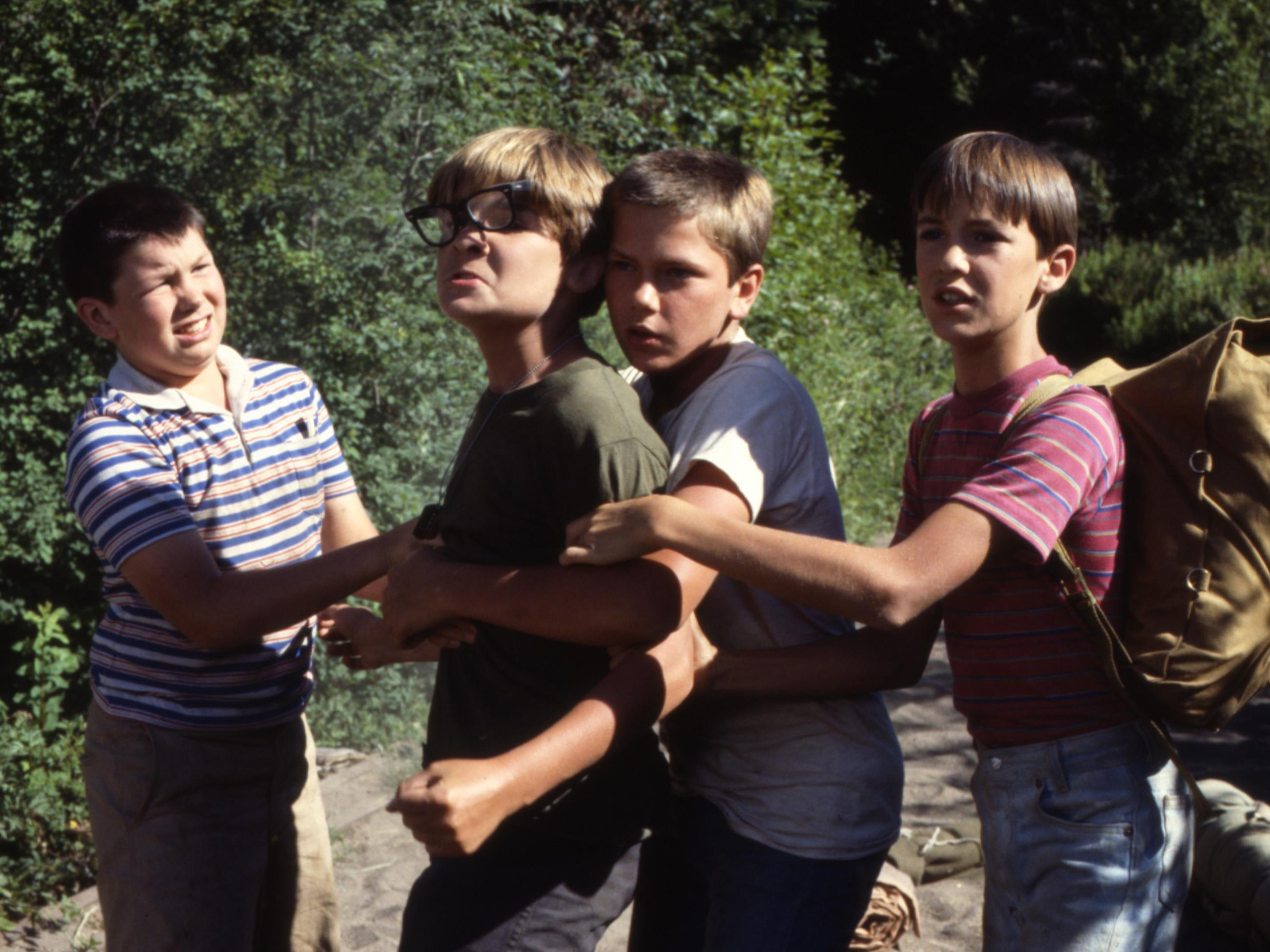 Four boys outdoors playing tug-of-war with rope, wearing striped and solid coloured shirts, dense green foliage in background.
