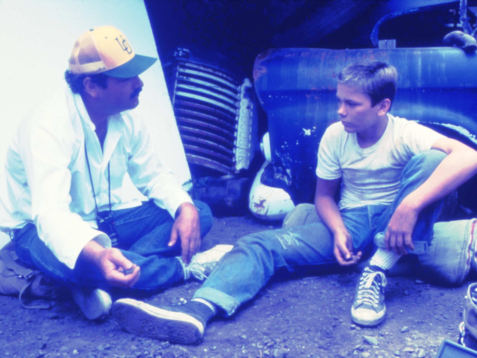 Two young men sitting on ground beside car, one wearing baseball cap and white shirt, other in grey t-shirt and trainers.