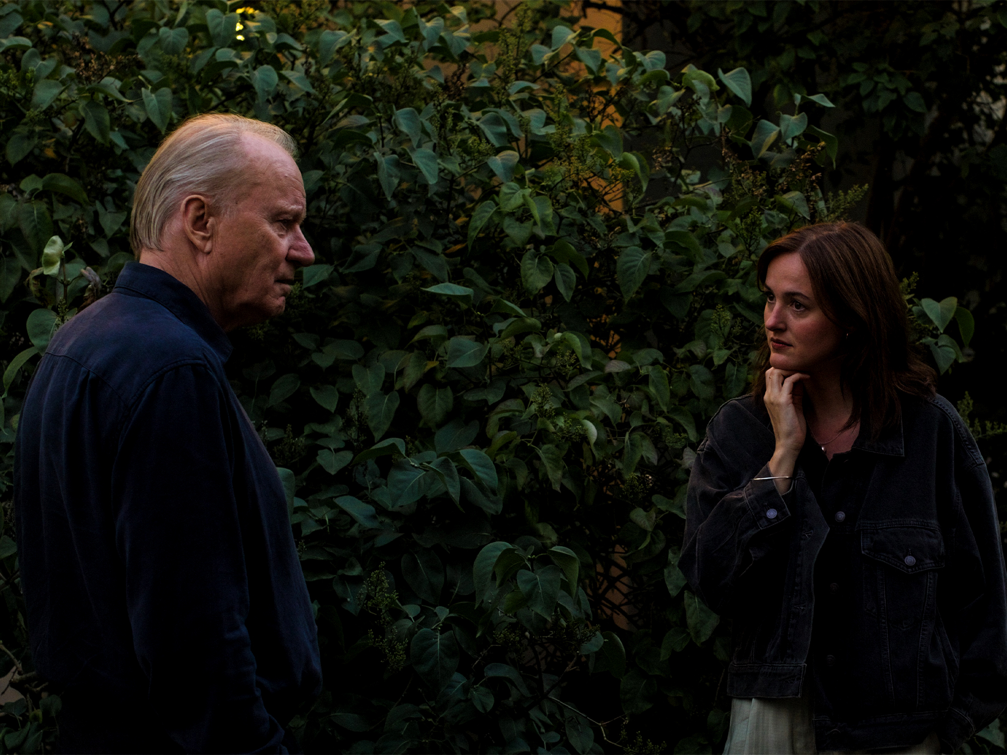 Older man in dark jacket speaking to younger woman with hand to chin, standing amongst dense green foliage.