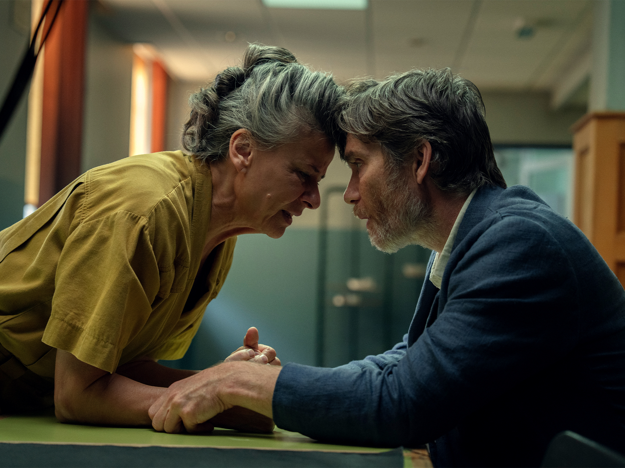 Two men with grey hair lean towards each other across green table, one in yellow shirt, other in dark blue jacket, institutional background.