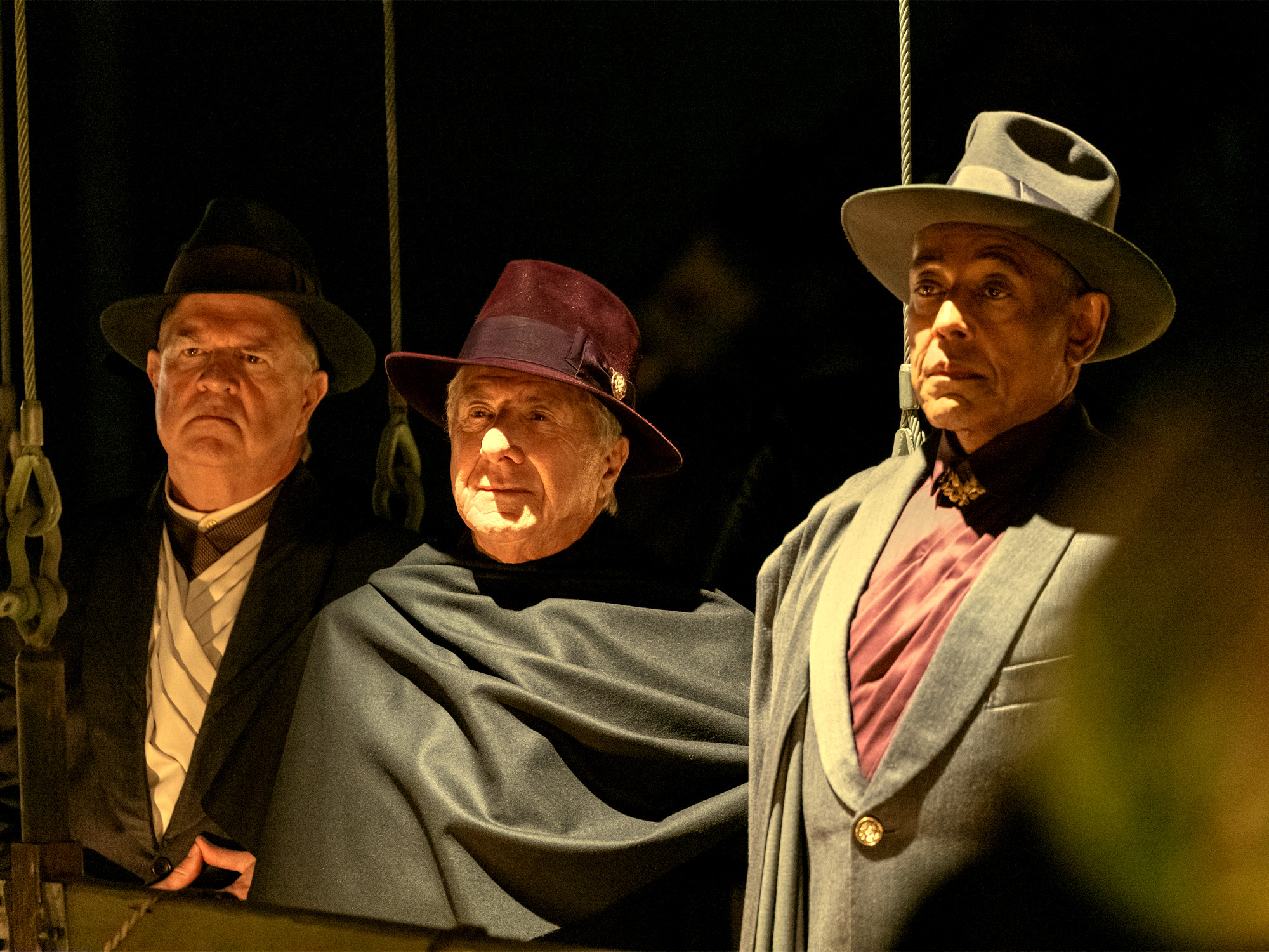 Three men in period costume on stage: one in dark suit with bowler hat, one in grey cape with burgundy hat, one in light suit with wide-brimmed hat.