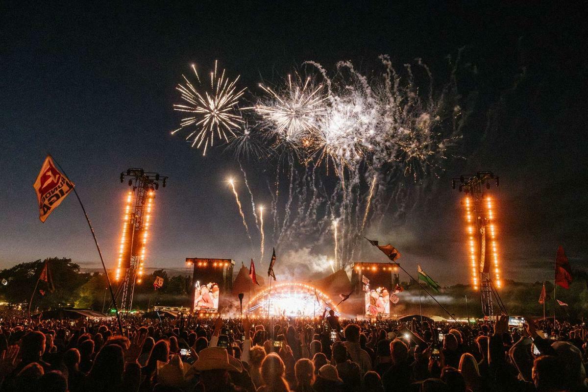 Fireworks exploding above outdoor music festival stage with crowd silhouettes, orange lighting towers, and dark evening sky.