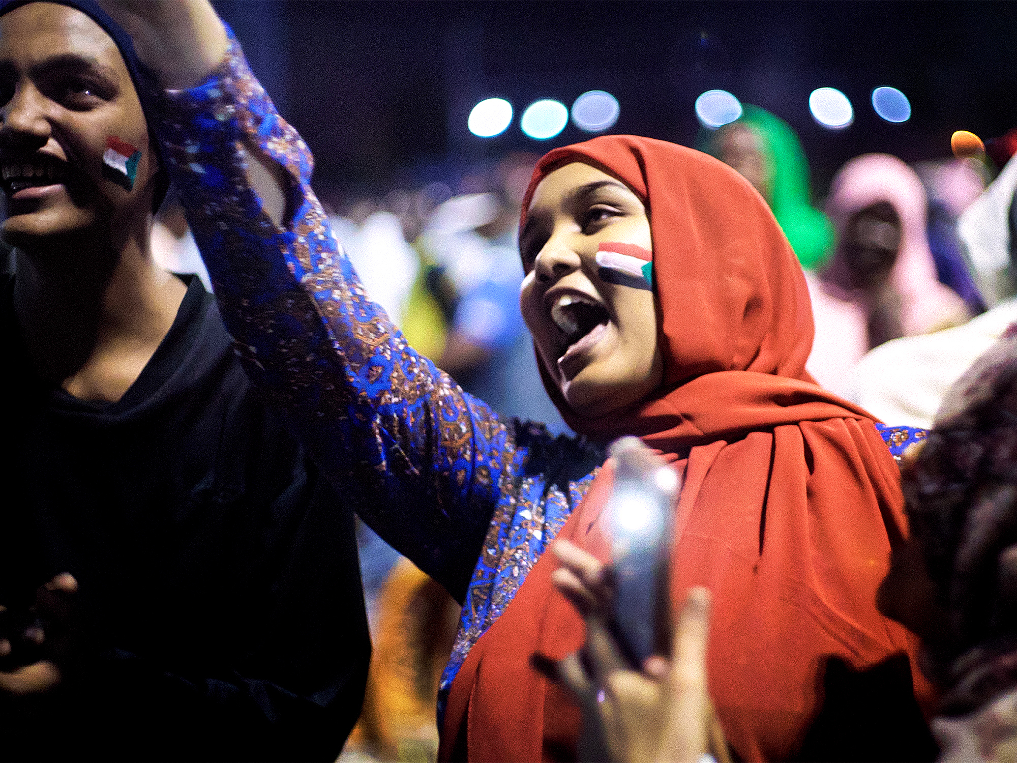 Two people with the Sudanese flag painted on their face at a night-time event, with colourful lights and a crowd visible in the background.
