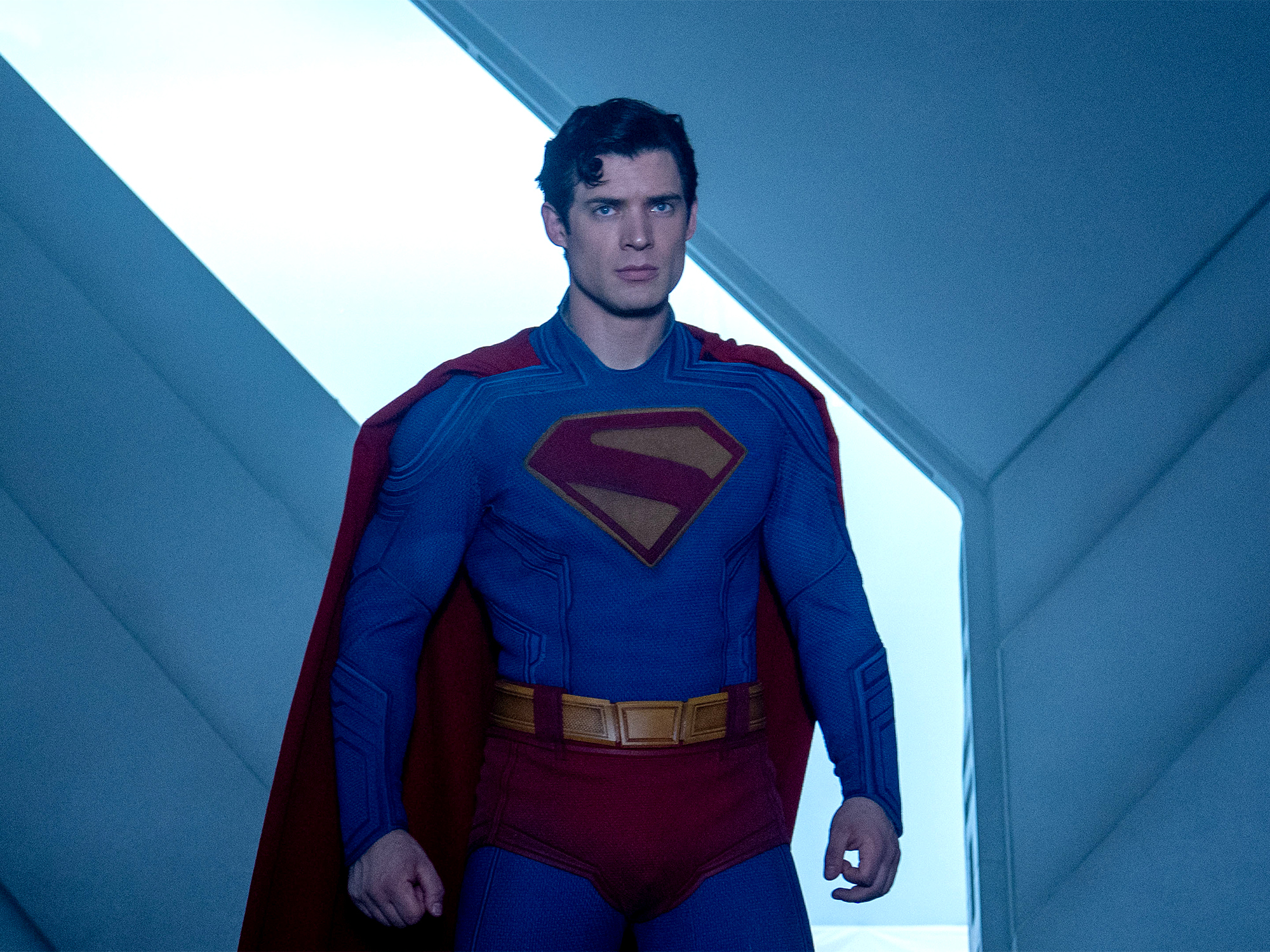 Man in blue Superman costume with red cape and "S" logo stands in white-lit corridor with angular grey walls.