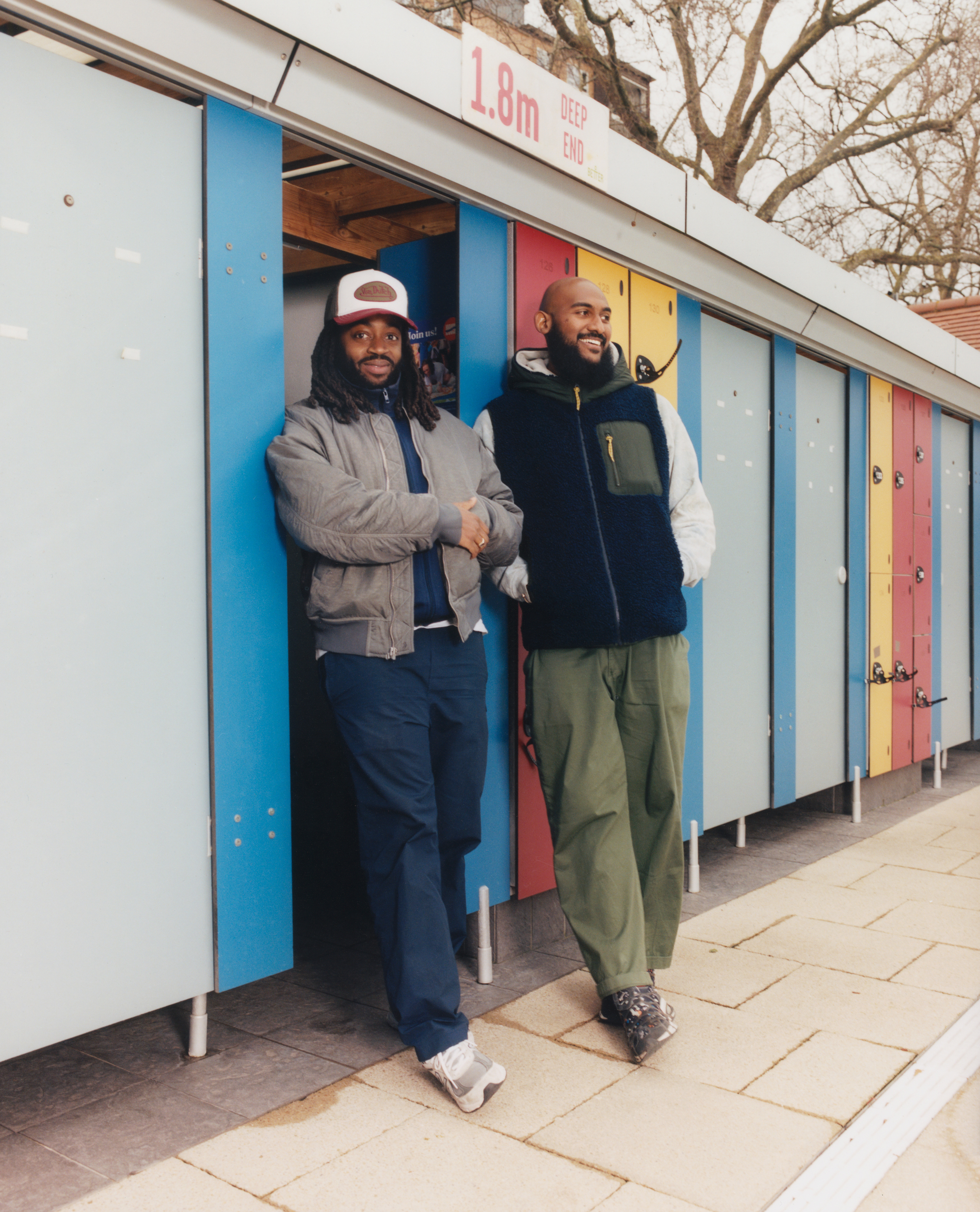 Two men standing outside colourful beach huts with blue, red, and yellow doors on concrete promenade under bare tree.