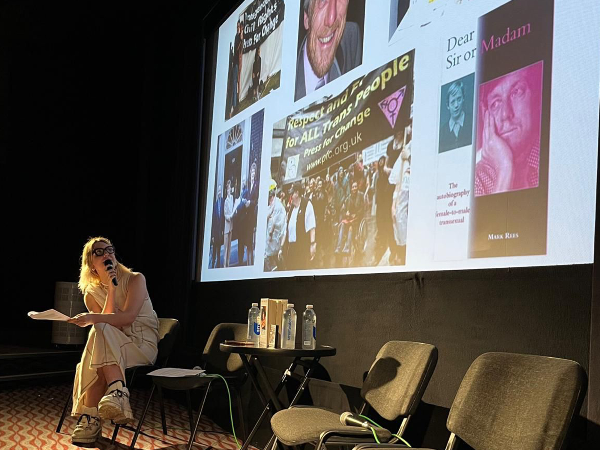 Projection screen displaying various book covers and magazine front pages; woman seated in foreground with book in hand.