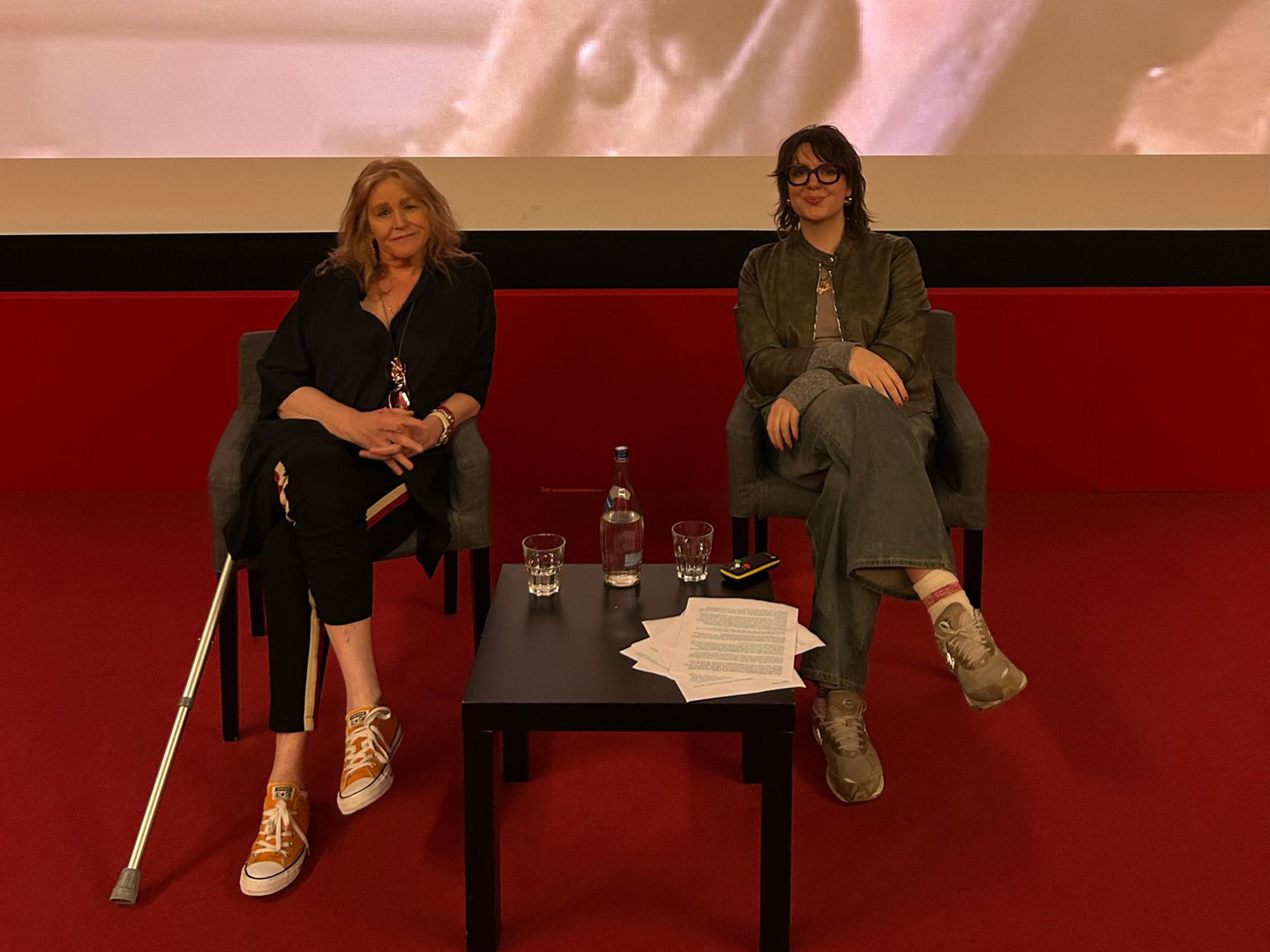 Two women seated on stage, one in a black outfit and one in a green jacket, engaged in discussion.