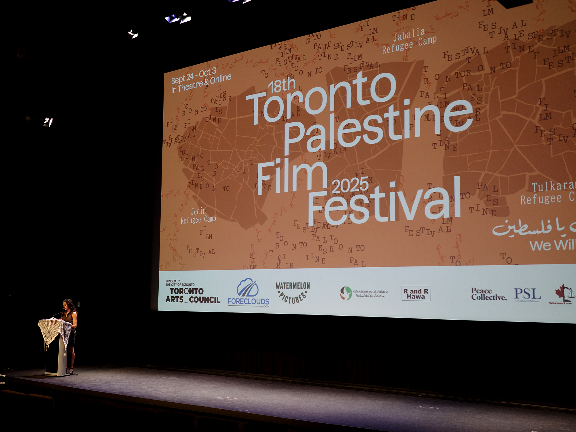 Speaker at podium on dark stage with large orange projection screen displaying "18th Toronto Palestine Film Festival 2025" text and sponsor logos.