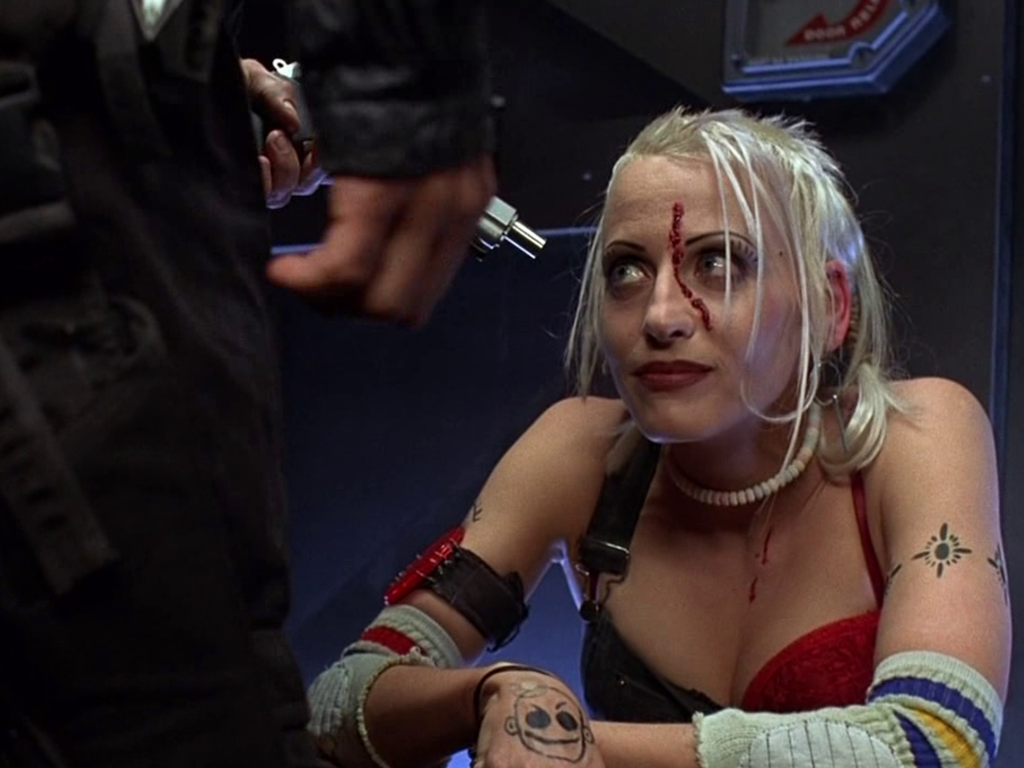 Lori Petty as Tank Girl, with blonde hair and blood dripping down her face, wearing a red top, looking at a hand brandishing a gun pointed at her.