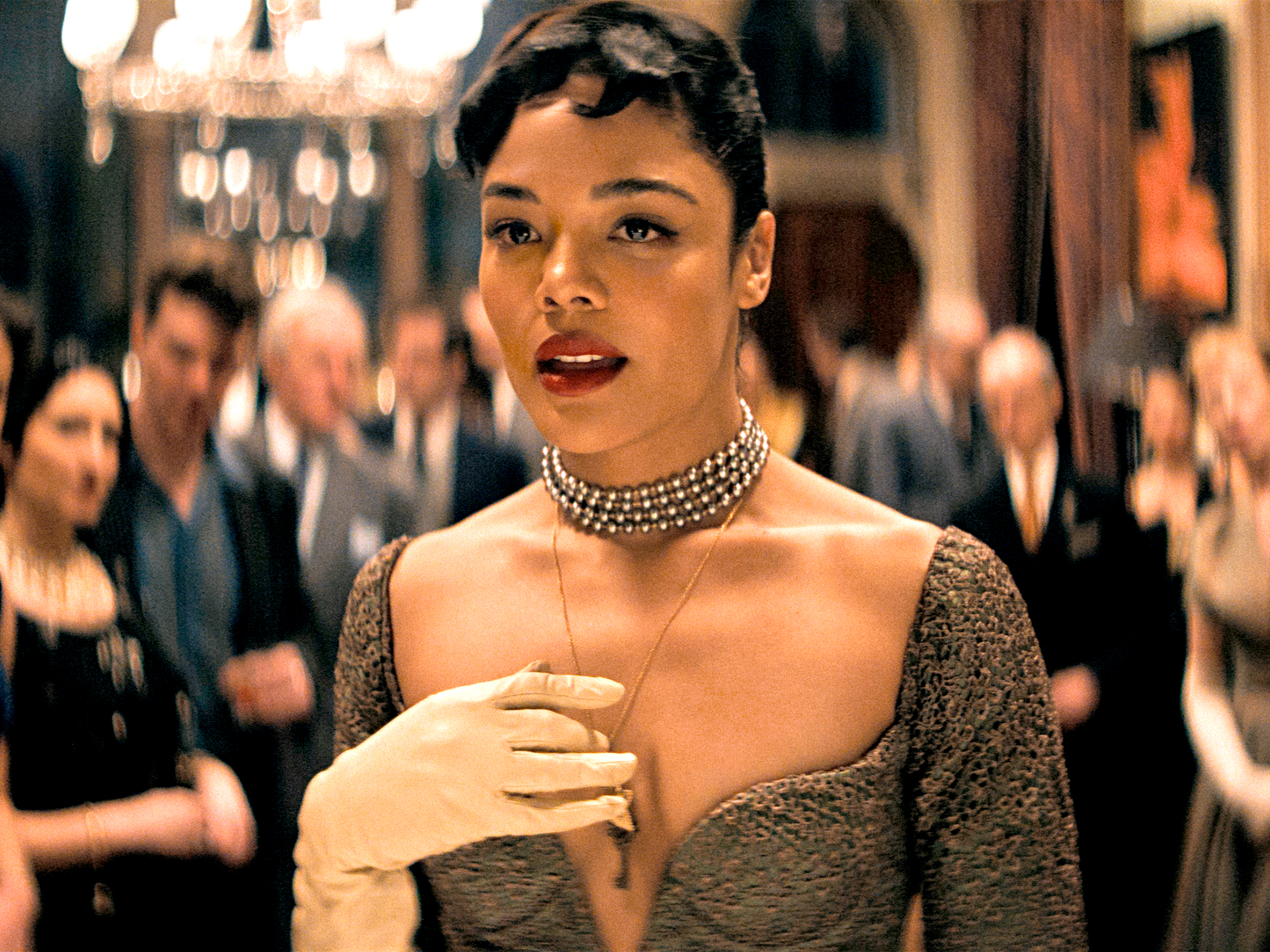 Woman in grey off-shoulder dress with pearl choker necklace at formal event with blurred crowd and chandelier in background.
