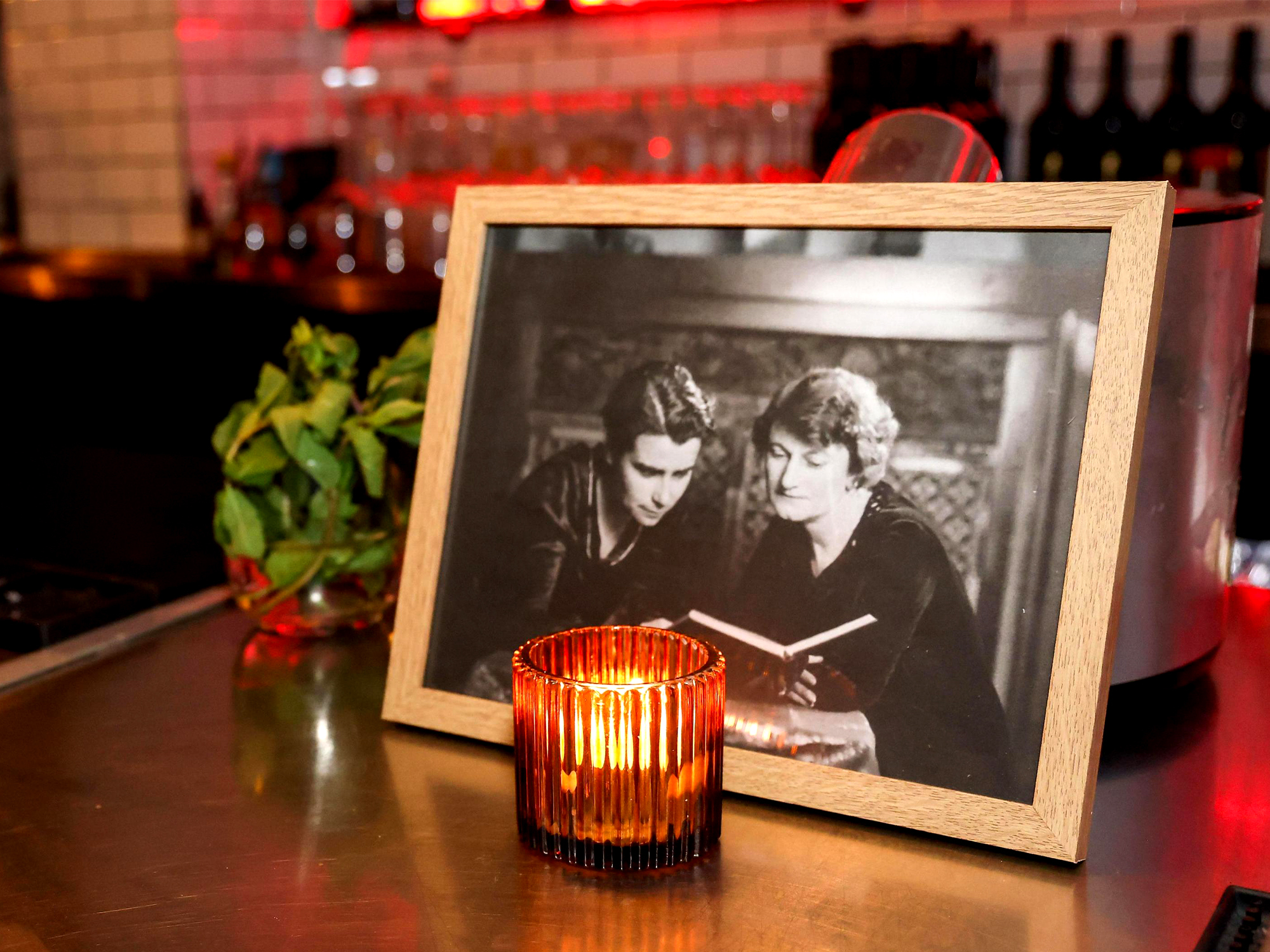 Framed black and white photograph of two people reading beside red glass candle holder on wooden table in dimly lit restaurant.