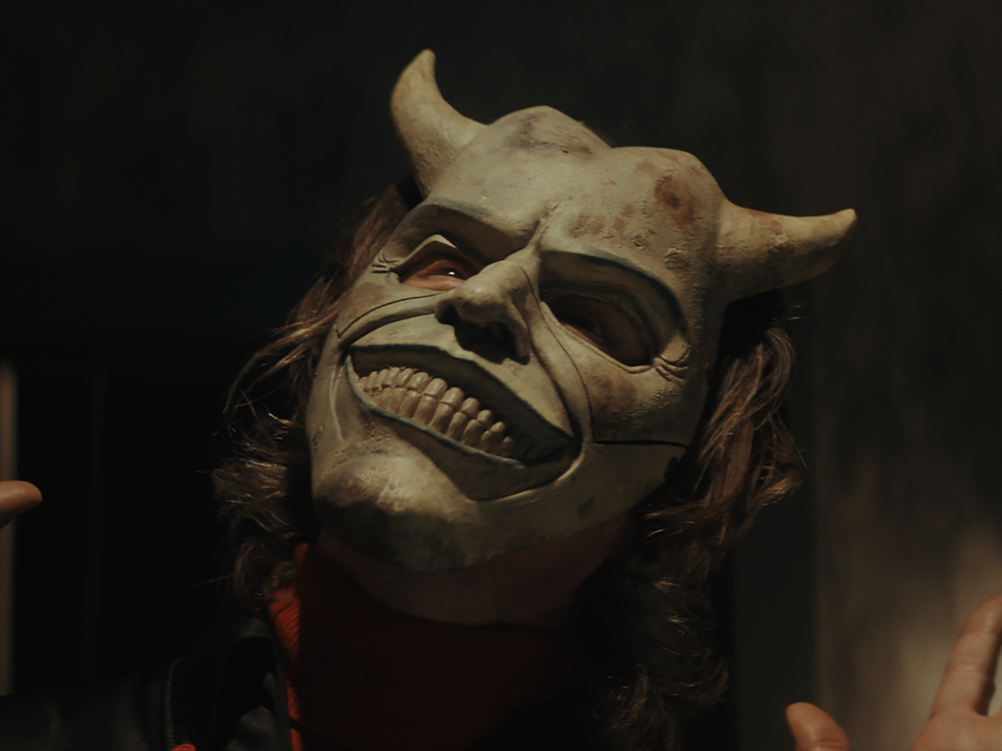 Eerie horned mask with grinning teeth and dark eyes on a dark background.