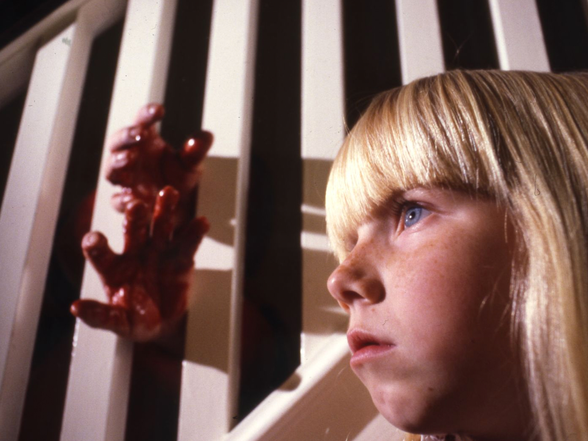 Blonde girl with blue eyes looking through bars, holding a red object.