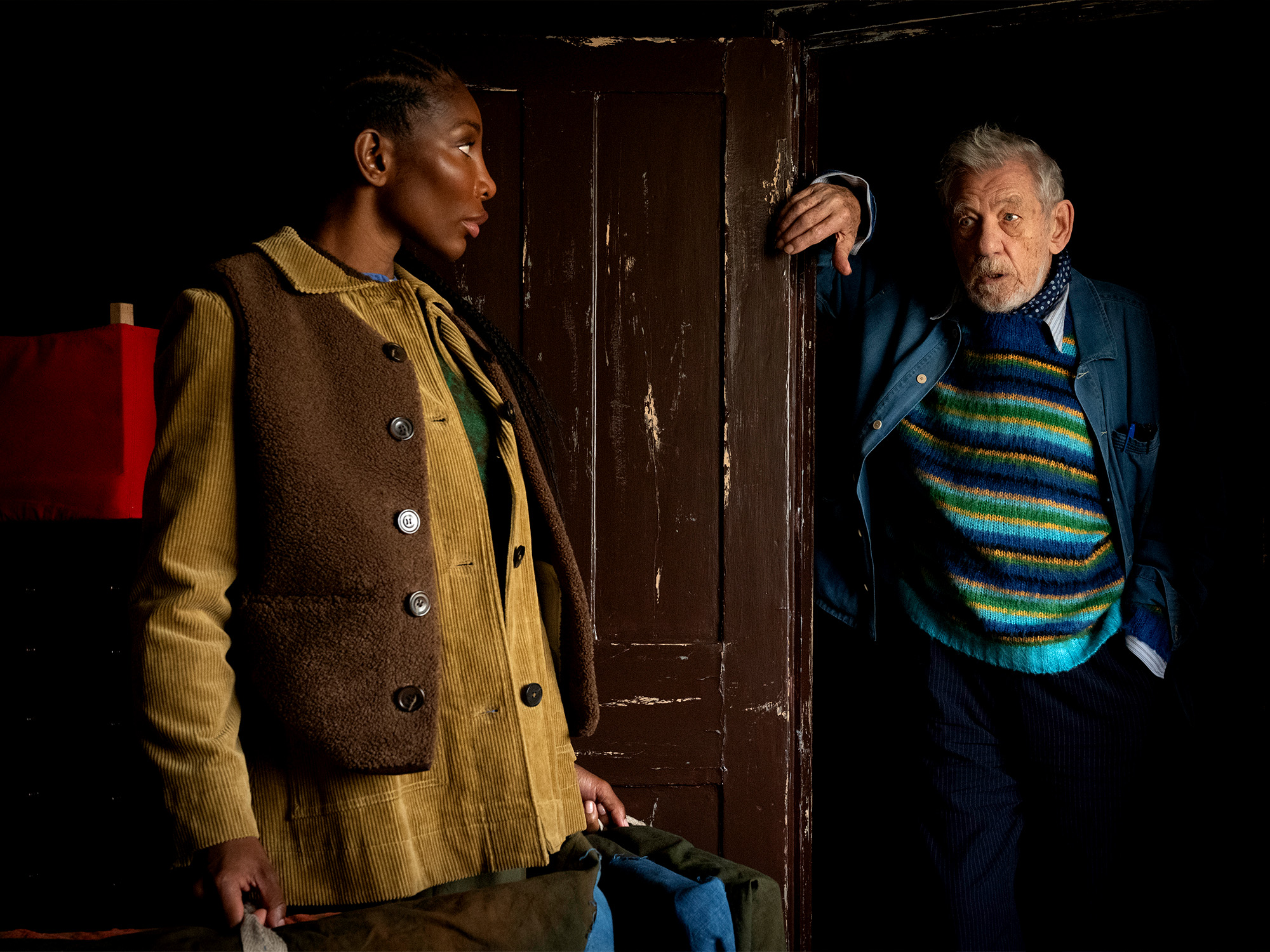 Black man in brown military-style coat stands beside wooden door, facing older white man in blue striped jumper in dimly lit interior.