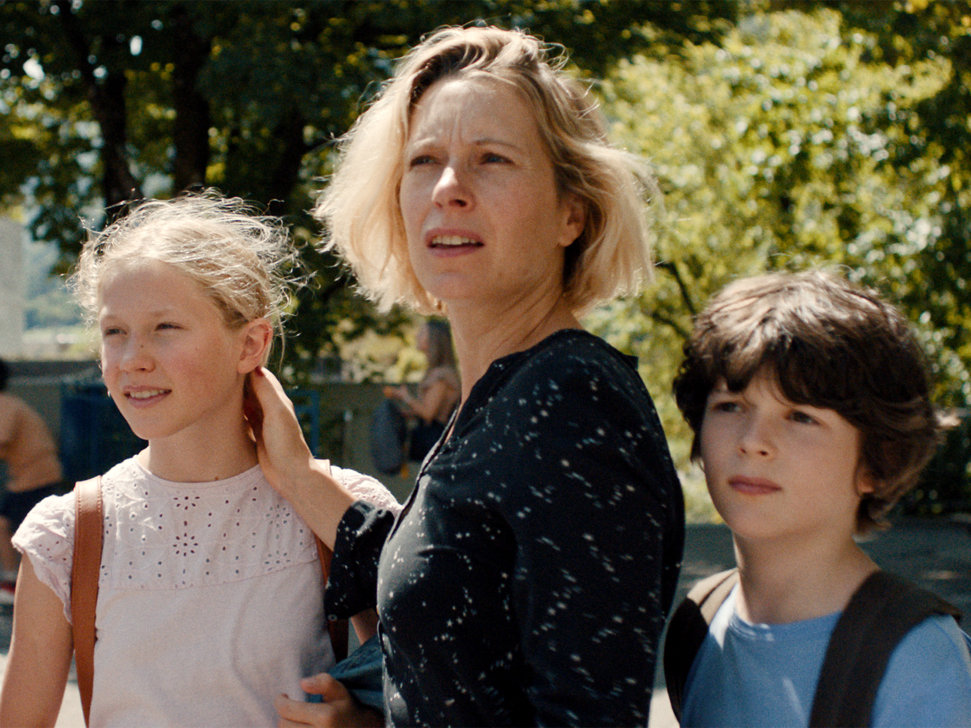 Woman with blonde bob haircut in dark top stands between two children - blonde girl in white top and boy with dark hair in blue shirt, outdoors with green foliage behind.