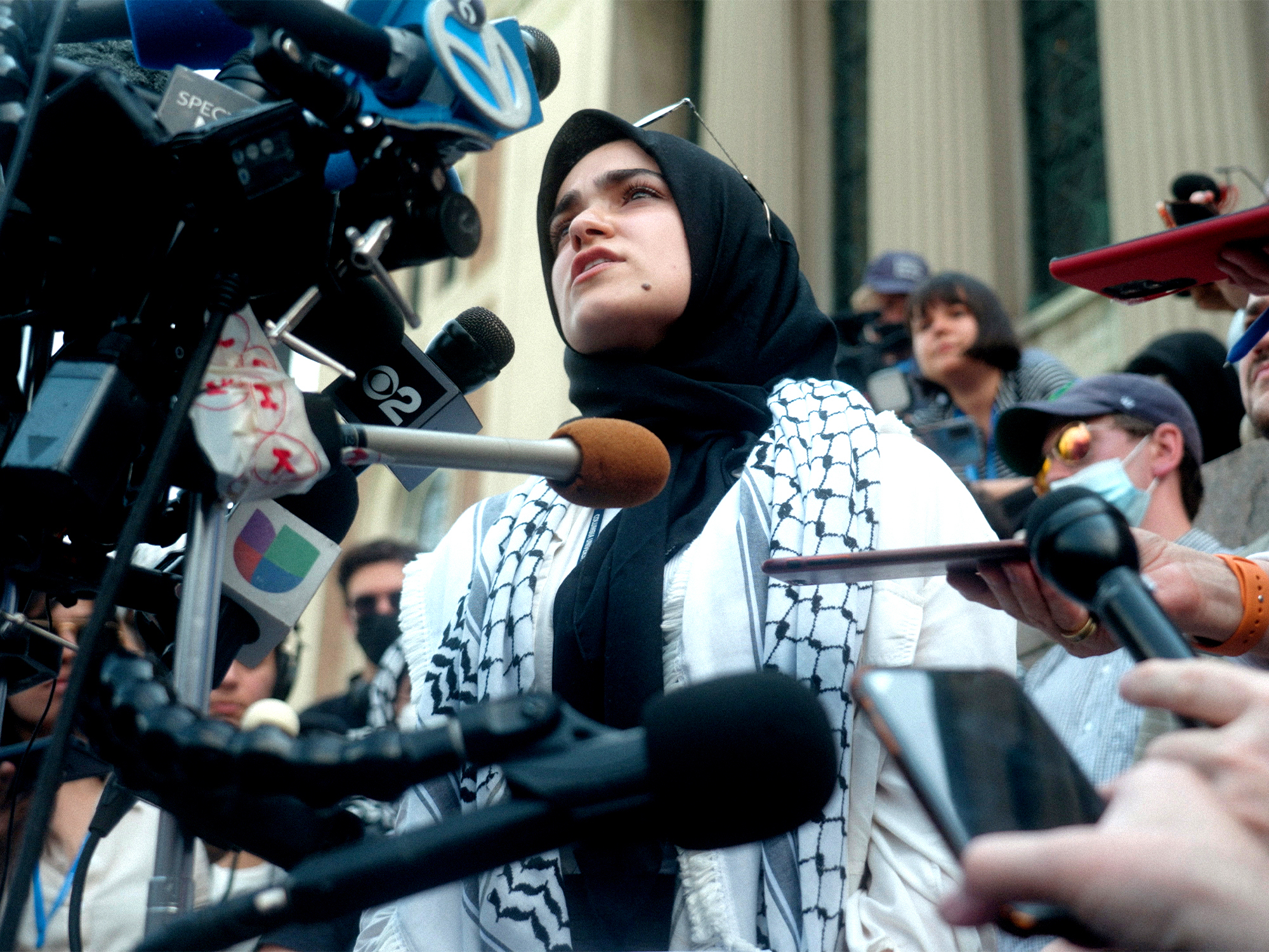 Woman in hijab and keffiyeh speaking into multiple microphones amidst crowd.