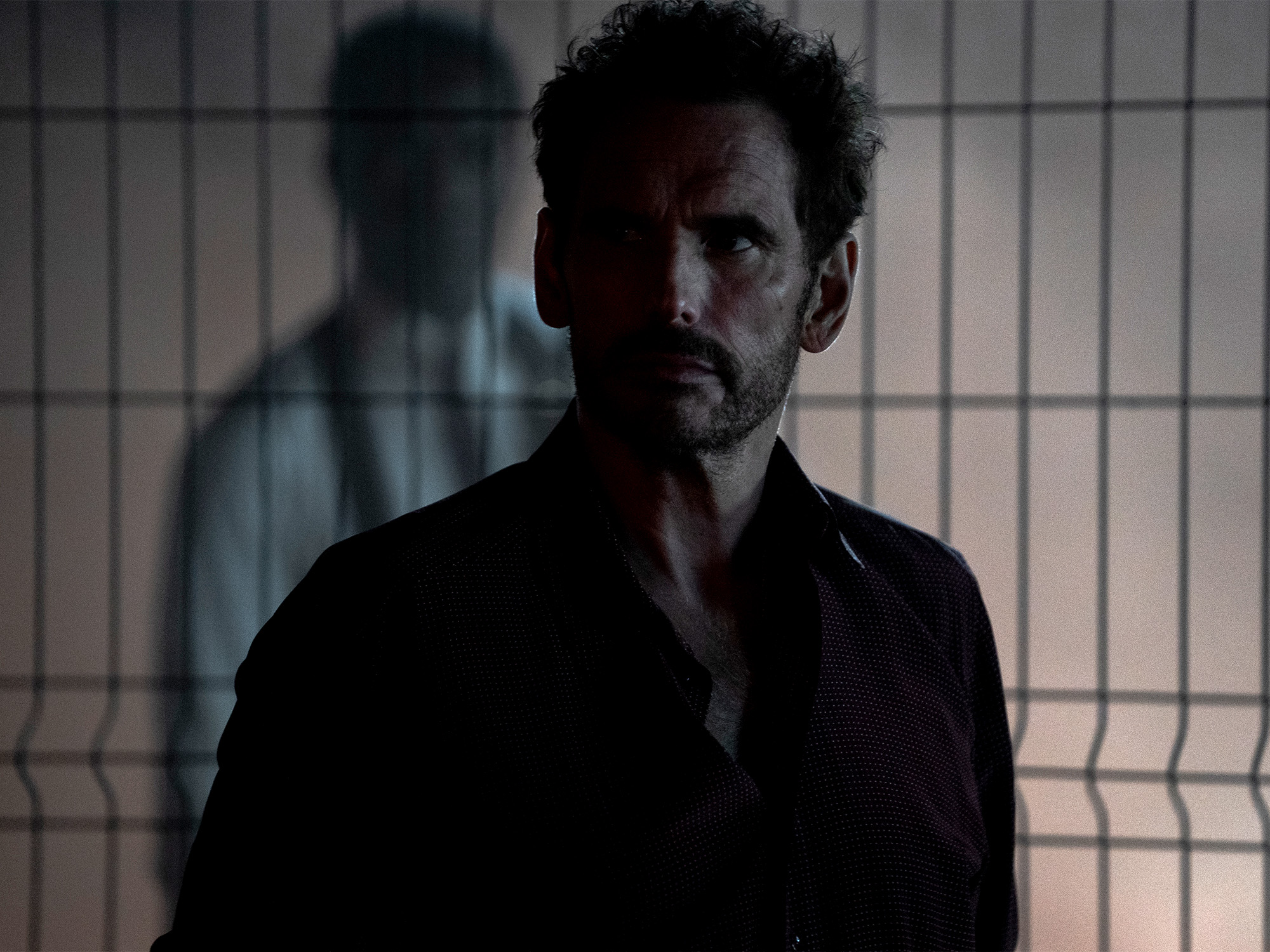 Man with curly hair and beard wearing dark shirt stands behind wire mesh fence, with blurred figure visible in background.