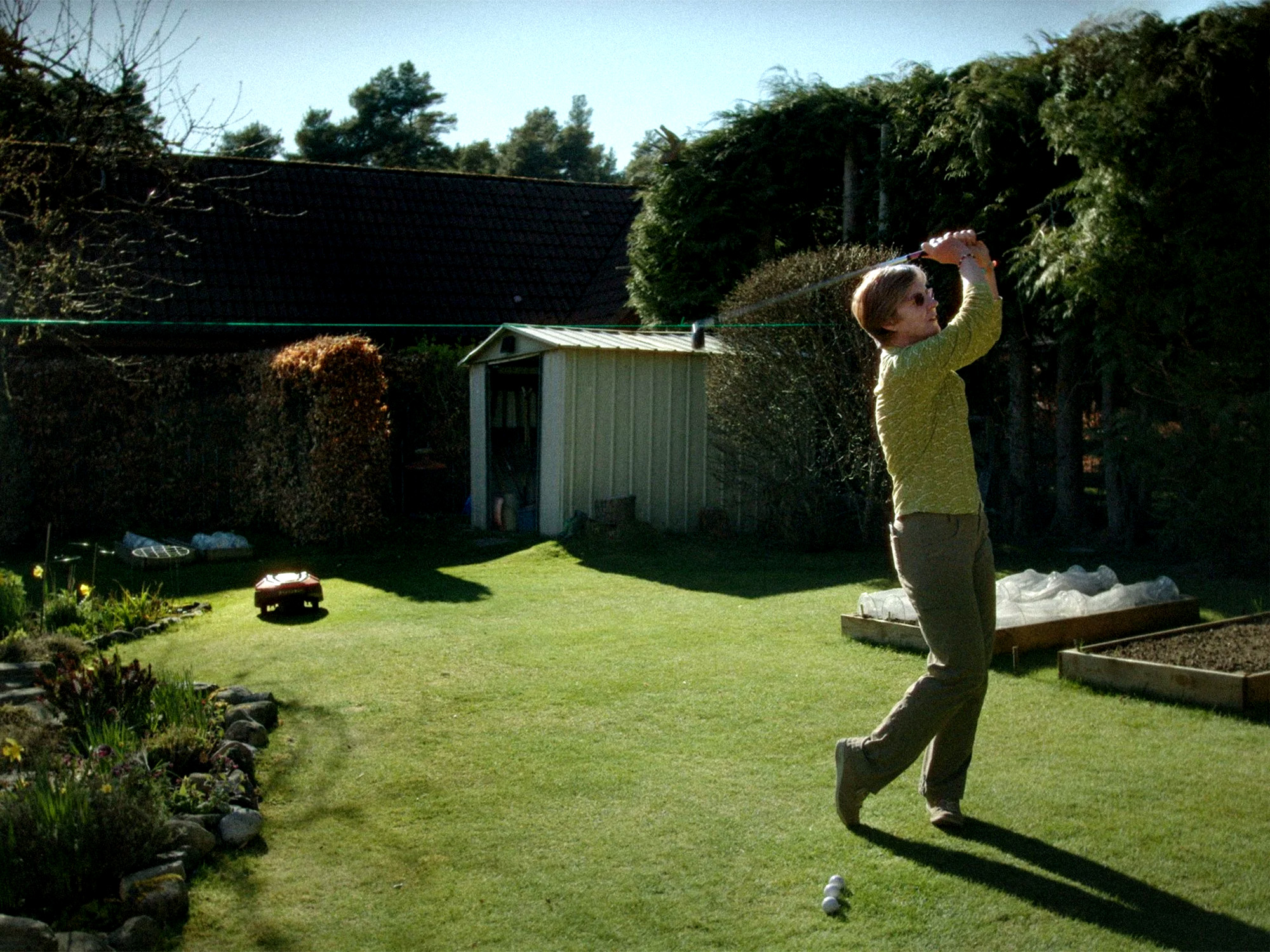 Man in yellow top mid-golf swing on lawn with green shed, hedge boundary, and garden borders in background.
