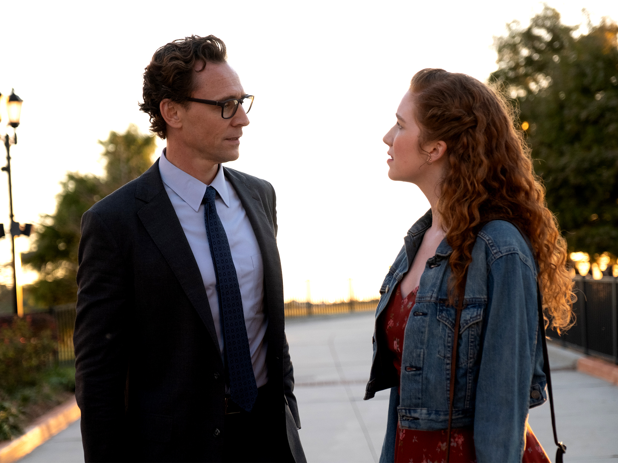 Man in dark suit and glasses faces woman in denim jacket with long curly hair on waterfront path at sunset.