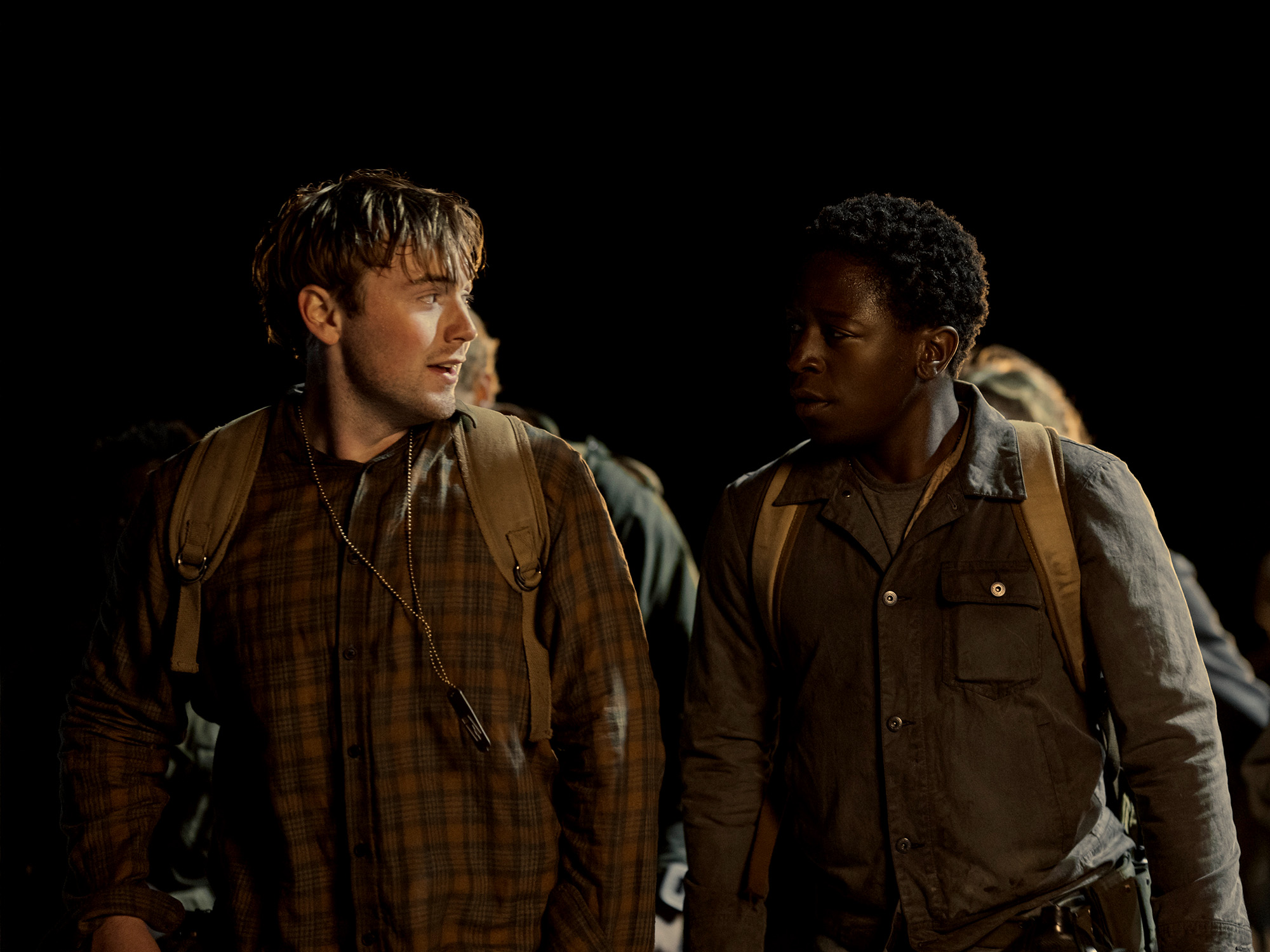 Two men in military uniforms against black background, one blonde looking left, one Black looking right, both wearing brown jackets.