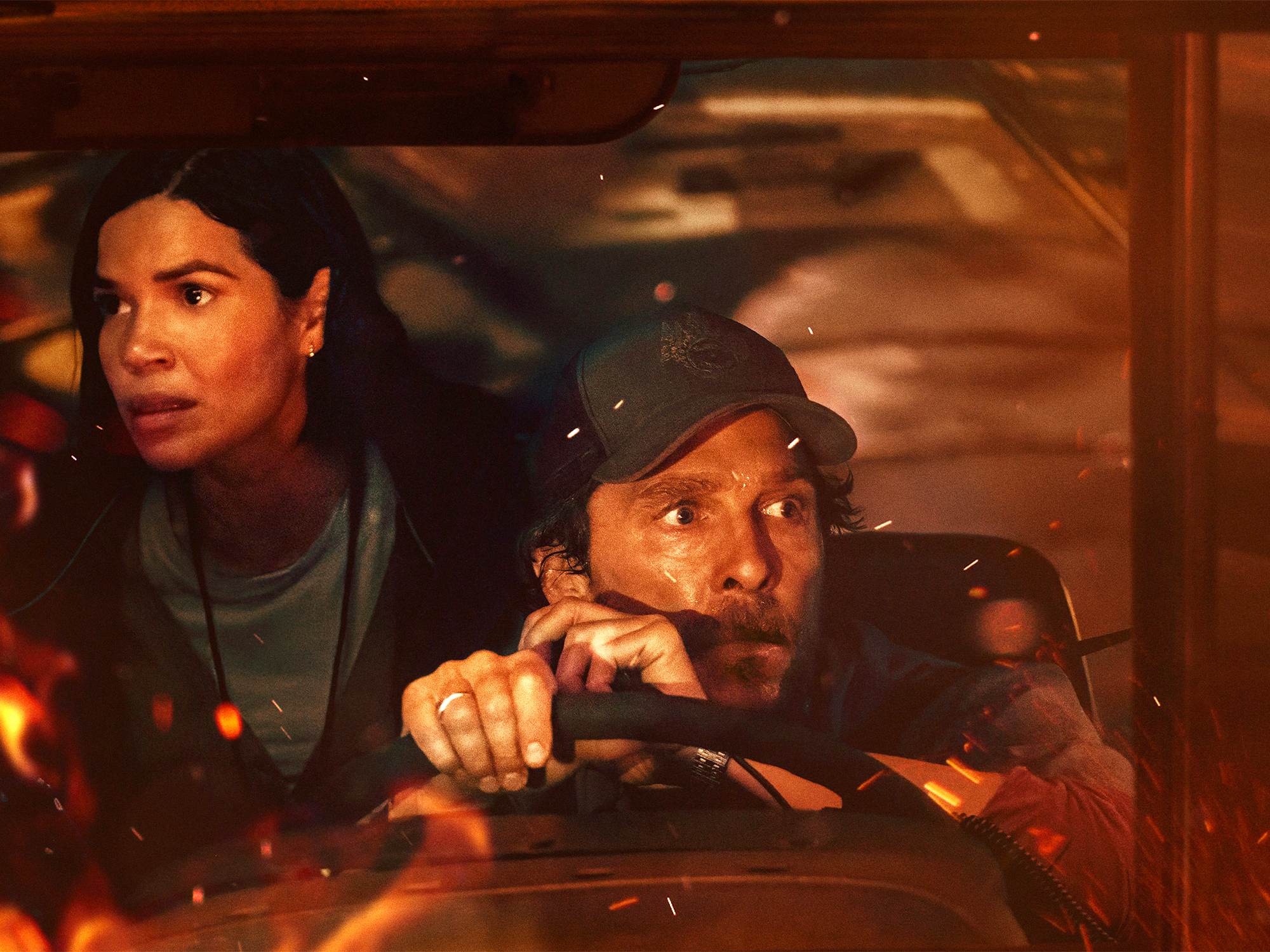 Two people inside a vehicle at night, man with beard and cap driving whilst speaking on phone, woman passenger visible on left.