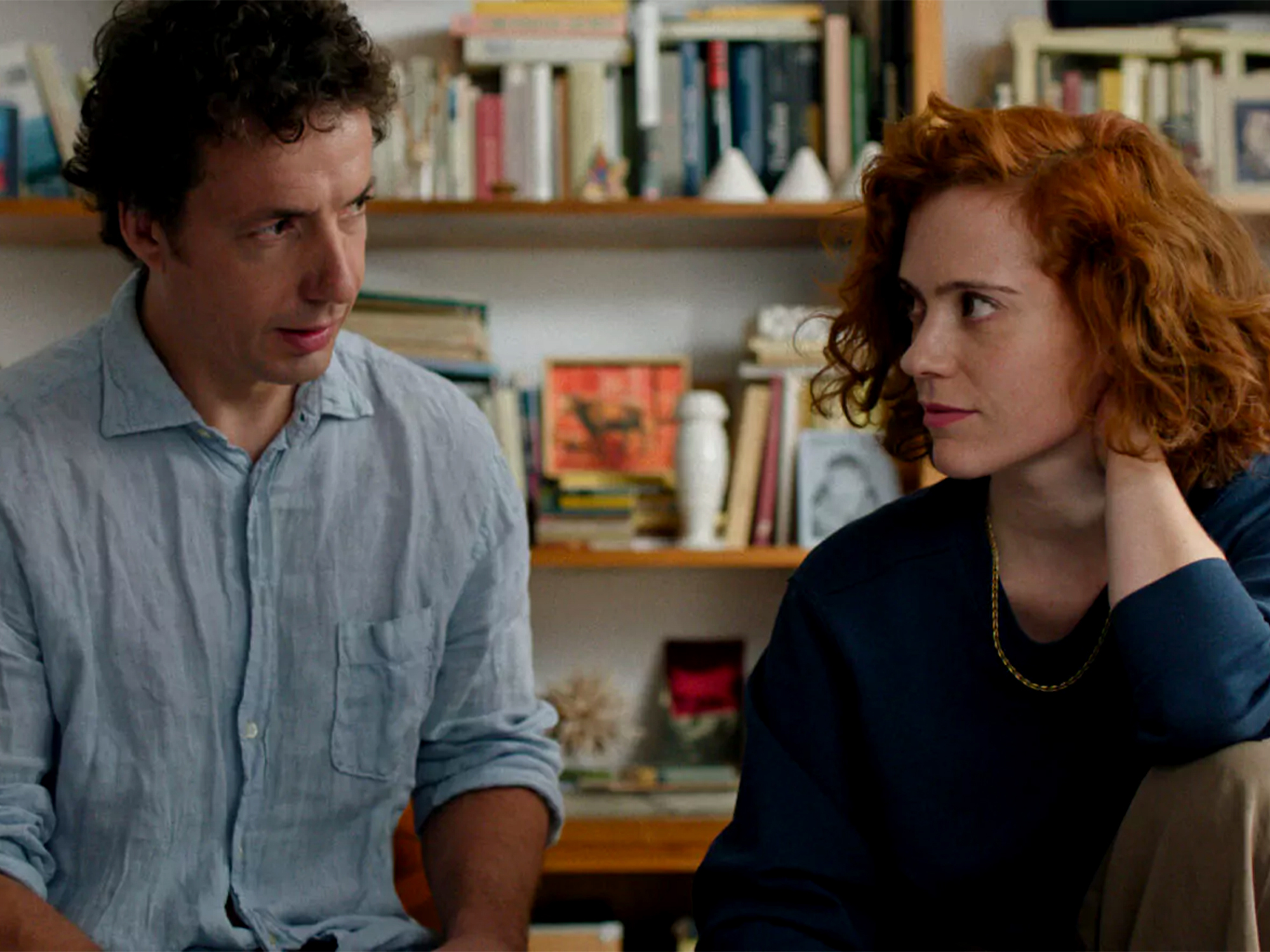Man in light blue shirt and woman with curly red hair in dark top having conversation in front of bookshelf filled with books.