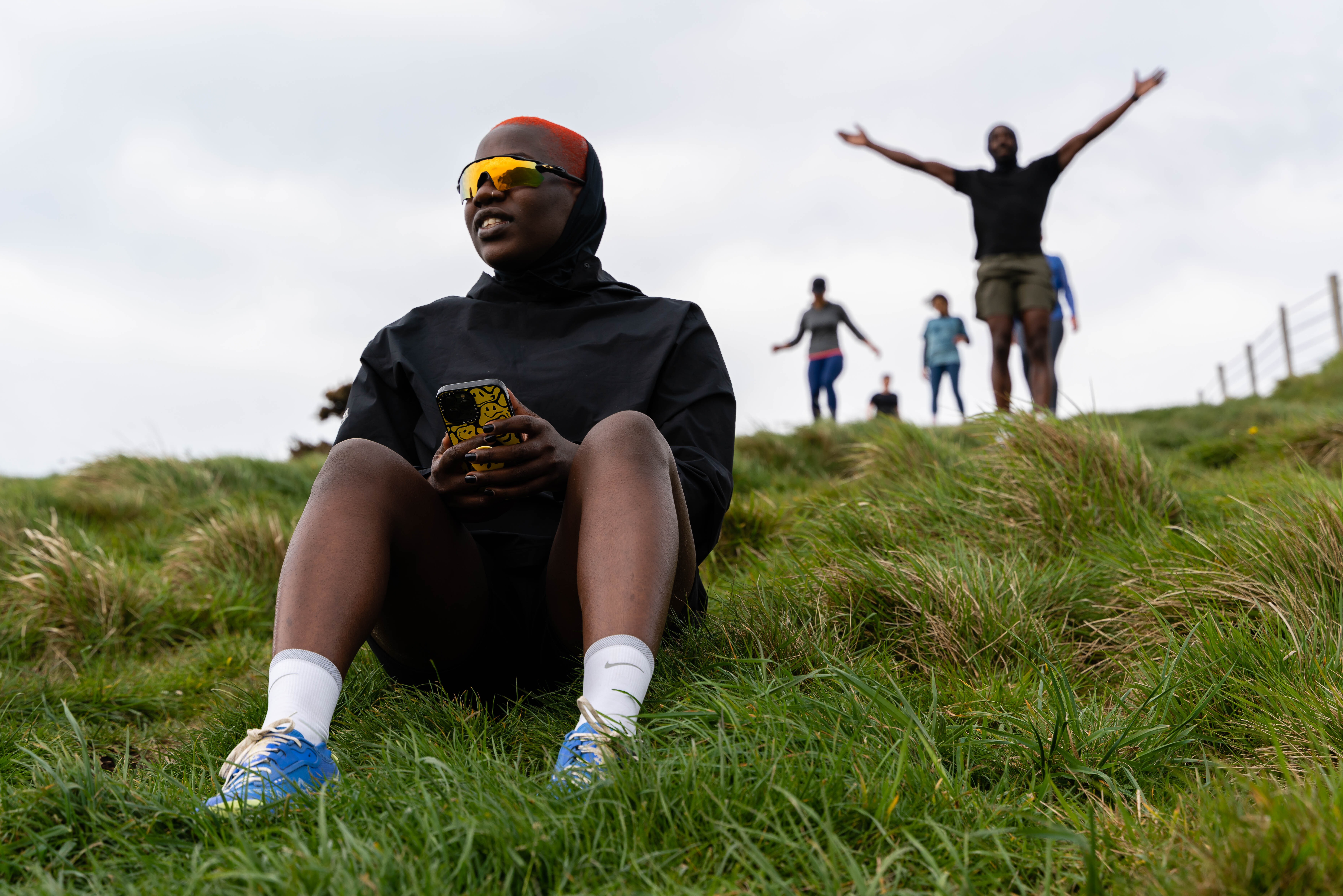 Person in black hoodie and yellow sunglasses sits on grassy hill holding phone, three people standing behind on hillside.