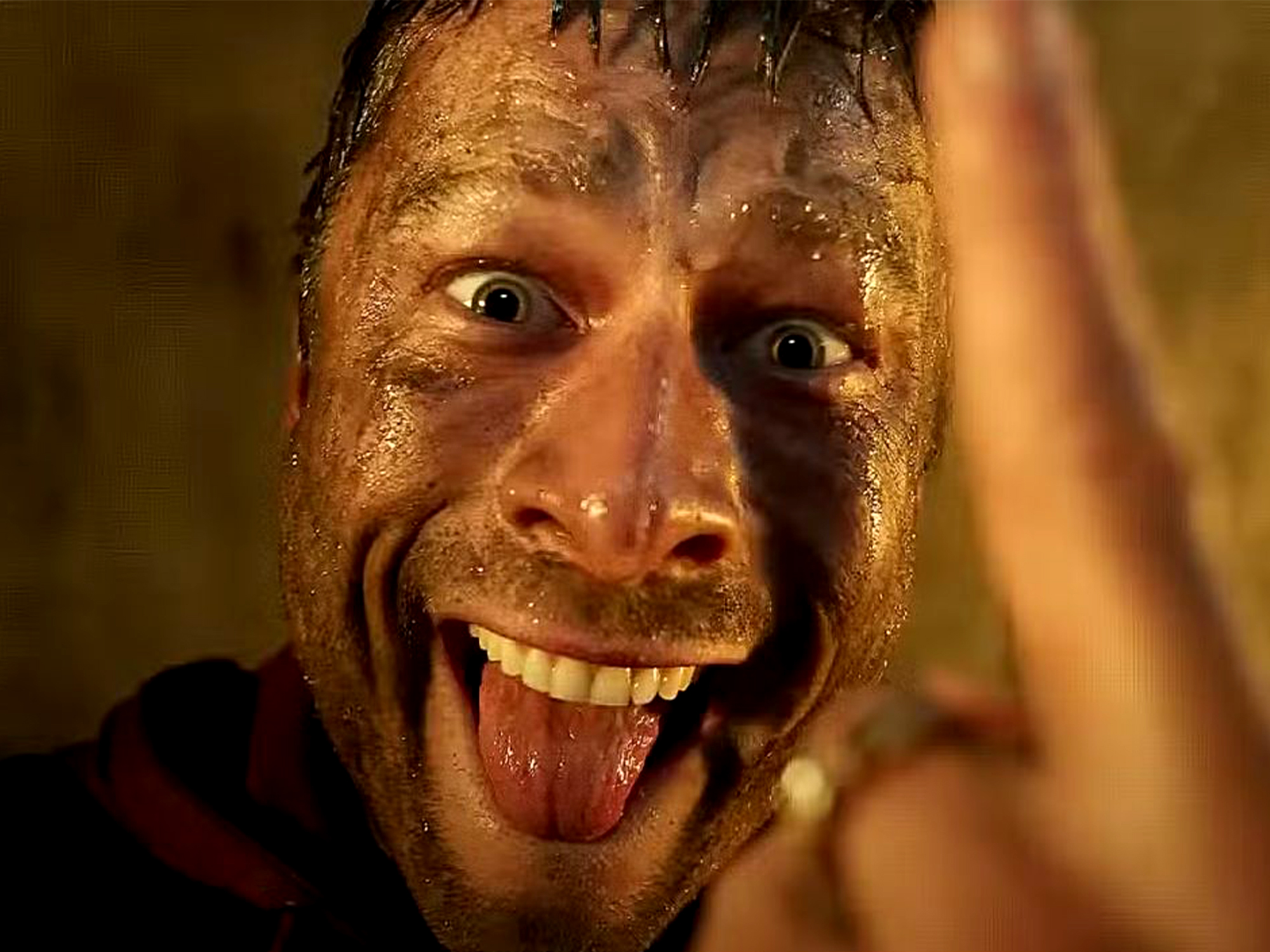Man with wet dark hair grinning widely, tongue out, hand raised towards camera. Face appears sweaty with warm golden lighting.