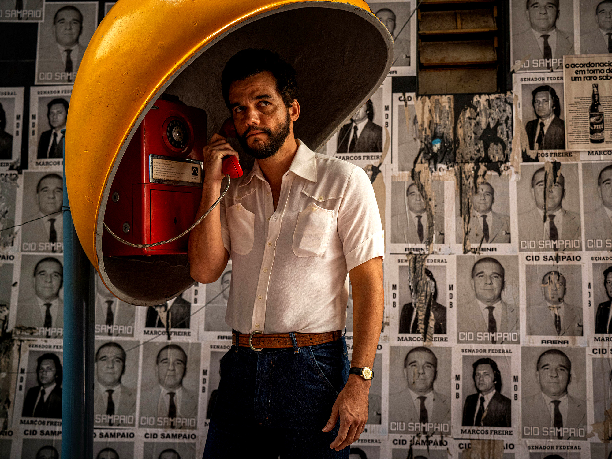 A man with a beard wearing a white shirt and holding a red telephone in front of a wall covered with old newspaper clippings.