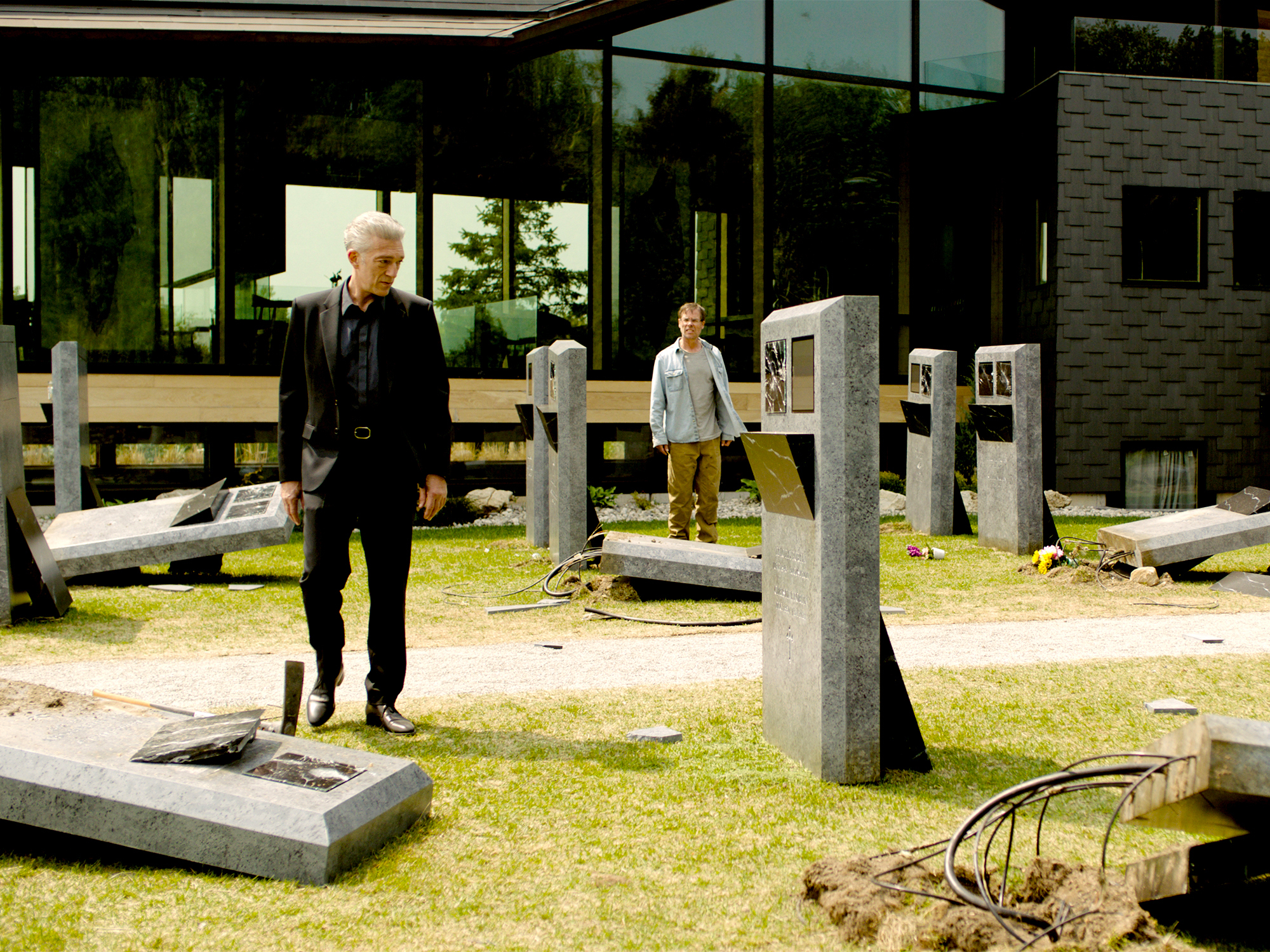 Two men standing amongst concrete construction materials and metal posts on grass with modern glass building in background.